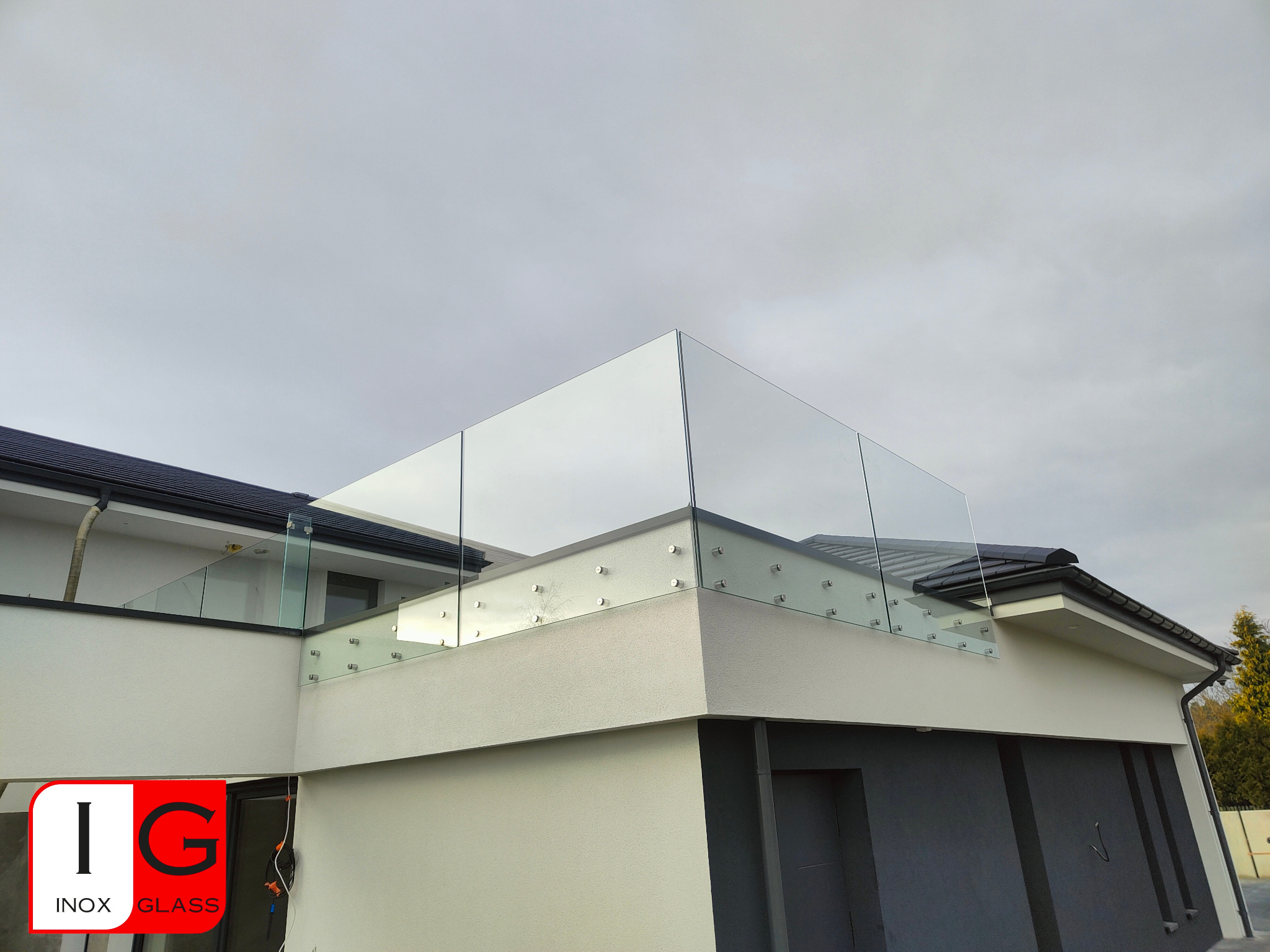 Nowoczesna, bezramowa konstrukcja ze szkła na balkonie, mocowana punktowo do ściany budynku o białej elewacji, widoczny fragment dachu z czarnej dachówki i logo firmy w lewym dolnym rogu.