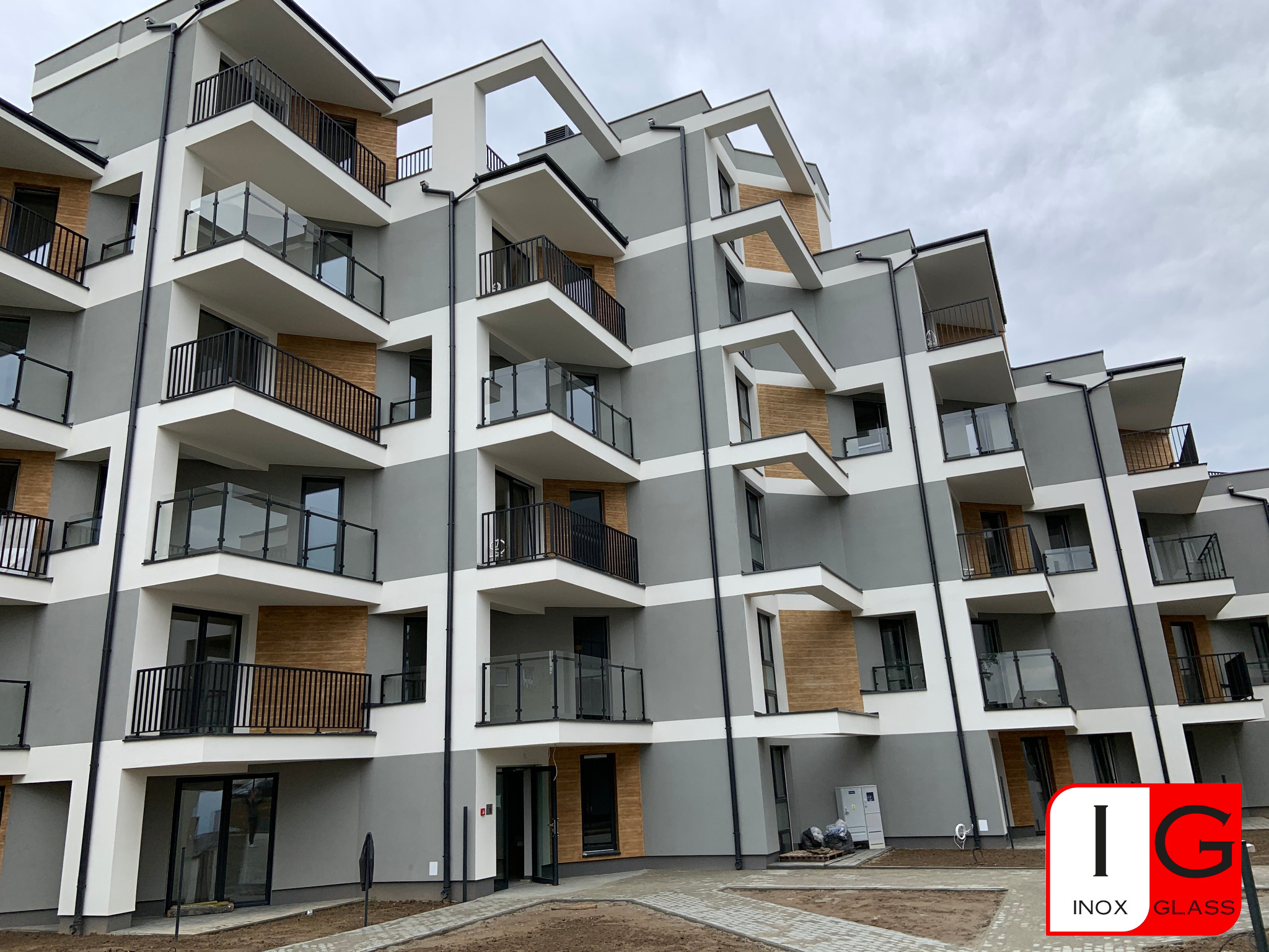 Nowoczesny budynek wielorodzinny z balkonami z balustradami szklanymi i metalowymi, widok z dołu, z logo firmy Inox Glass w prawym dolnym rogu.