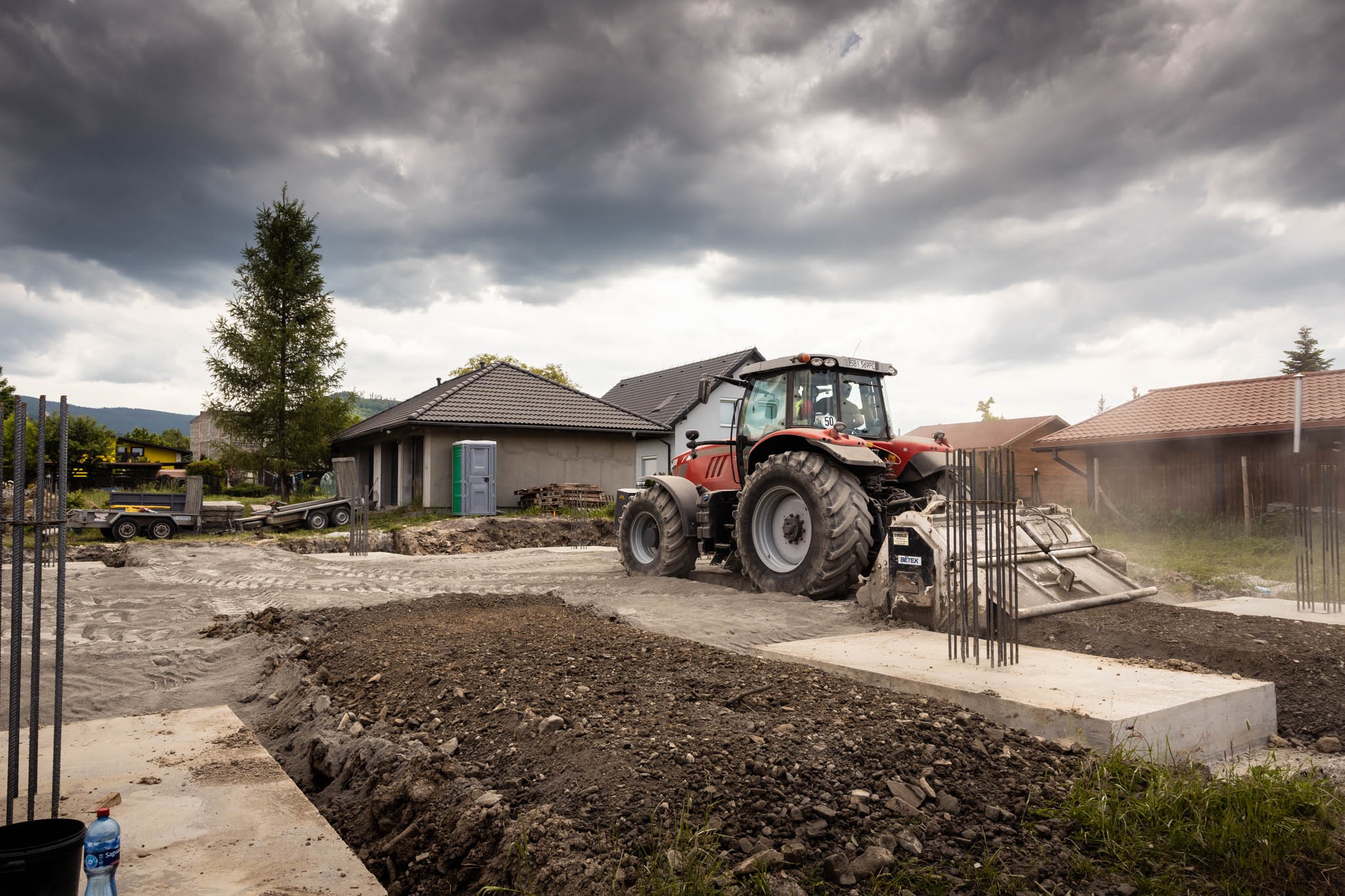 Czerwony traktor z równiarką wyrównuje teren pod budowę domu jednorodzinnego, widoczne fundamenty, zbrojenia i przenośna toaleta, pochmurne niebo w tle.