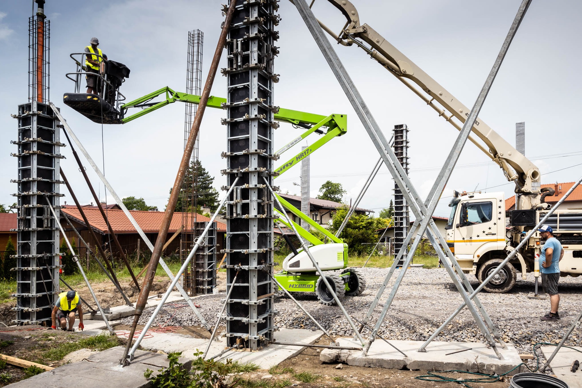 Wznoszenie żelbetowych słupów na placu budowy z użyciem szalunków systemowych, podnośnika koszowego i pompy do betonu, widoczni pracownicy w kamizelkach odblaskowych.