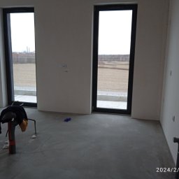 MAISTERBUD MYKHAILO KHAPKO - Surowe wnętrze z dwoma oknami, widoczne ślady po pracach wykończeniowych na ścianach i podłodze, narzędzia i ubrania robocze na podłodze, widok na zewnątrz przez okna.