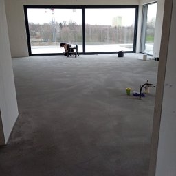 MAISTERBUD MYKHAILO KHAPKO - Kobieta na drabince czyści duże okno w surowym, niezamieszkałym wnętrzu z betonową podłogą. Widoczne wiadro, środki czystości i panorama za oknem.