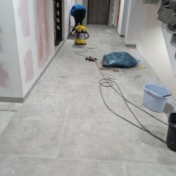 MAISTERBUD MYKHAILO KHAPKO - Korytarz w trakcie prac wykończeniowych: ściany częściowo zagruntowane, na podłodze płytki, widoczny odkurzacz przemysłowy, worek na śmieci, wiadro i szlifierka do gipsu.