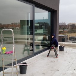 MAISTERBUD MYKHAILO KHAPKO - Pracownica w granatowej bluzie z kapturem siedzi na rozkładanej drabince podczas mycia okien w nowoczesnym budynku z dużymi przeszkleniami. Na pierwszym planie drabina z kubkiem i różową ścierką...