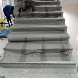 MAISTERBUD MYKHAILO KHAPKO - Granitowe schody zabezpieczone folią ochronną podczas prac remontowych, widoczna osoba w niebieskiej bluzie sprzątająca lub przygotowująca do dalszych prac, z odkurzaczem przemysłowym w tle.