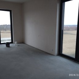 MAISTERBUD MYKHAILO KHAPKO - Pusty pokój w trakcie wykańczania, z drabiną, wiadrem i rolką papieru, ściany przygotowane do malowania, duże okna z widokiem na pole.