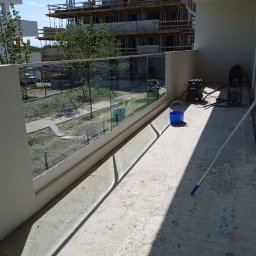 MAISTERBUD MYKHAILO KHAPKO - Betonowy balkon z szklaną balustradą w trakcie sprzątania, widoczny odkurzacz przemysłowy, niebieskie wiadro i butelka wody mineralnej, w tle budowa.