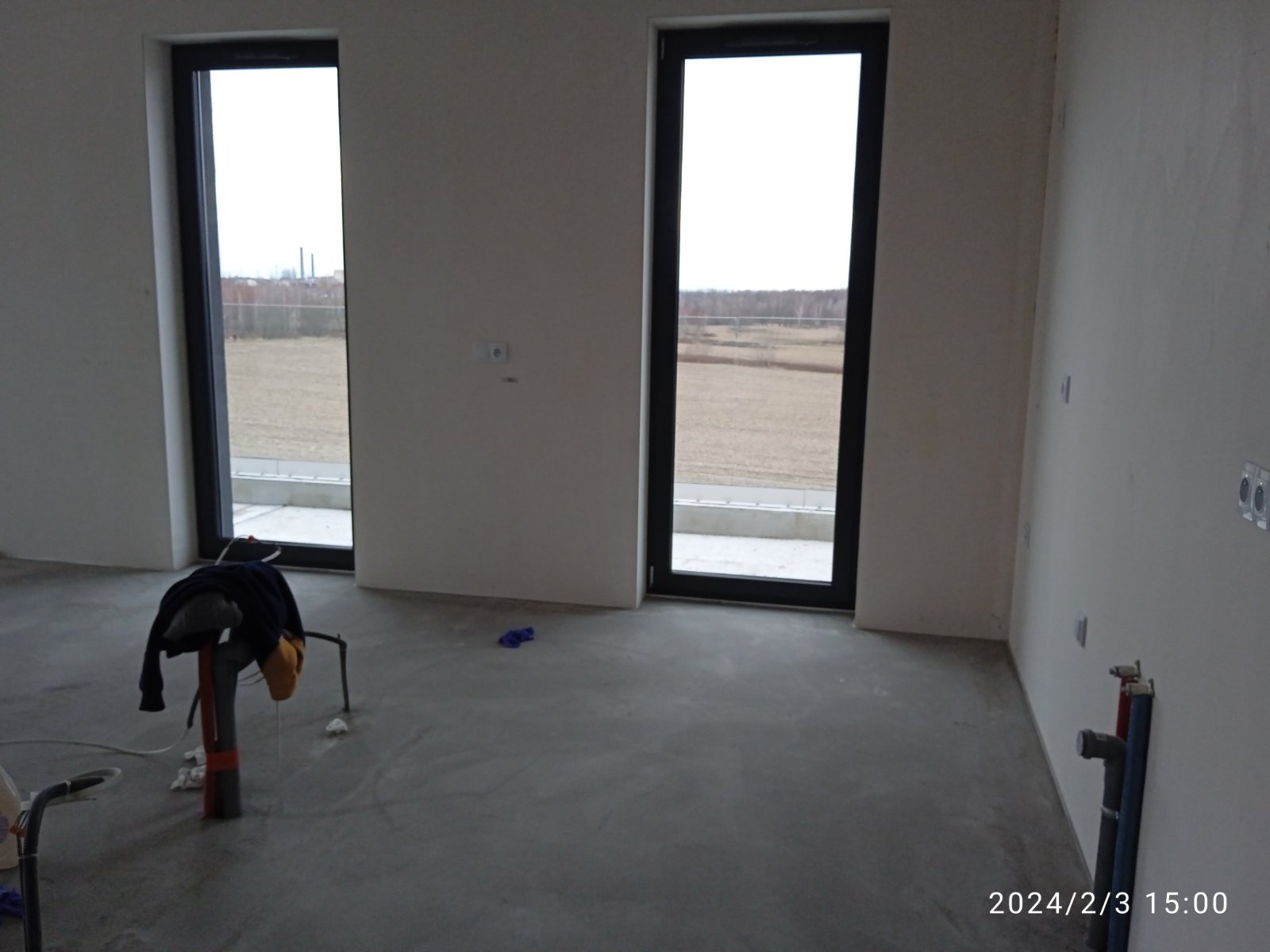 Surowe wnętrze z dwoma oknami, widoczne ślady po pracach wykończeniowych na ścianach i podłodze, narzędzia i ubrania robocze na podłodze, widok na zewnątrz przez okna.