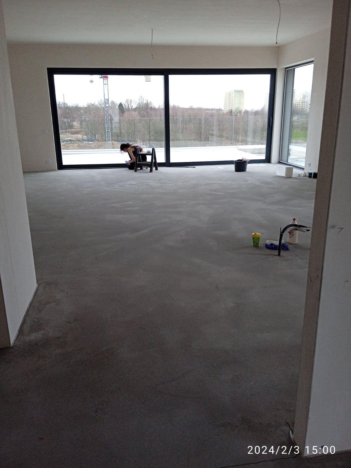 Kobieta na drabince czyści duże okno w surowym, niezamieszkałym wnętrzu z betonową podłogą. Widoczne wiadro, środki czystości i panorama za oknem.