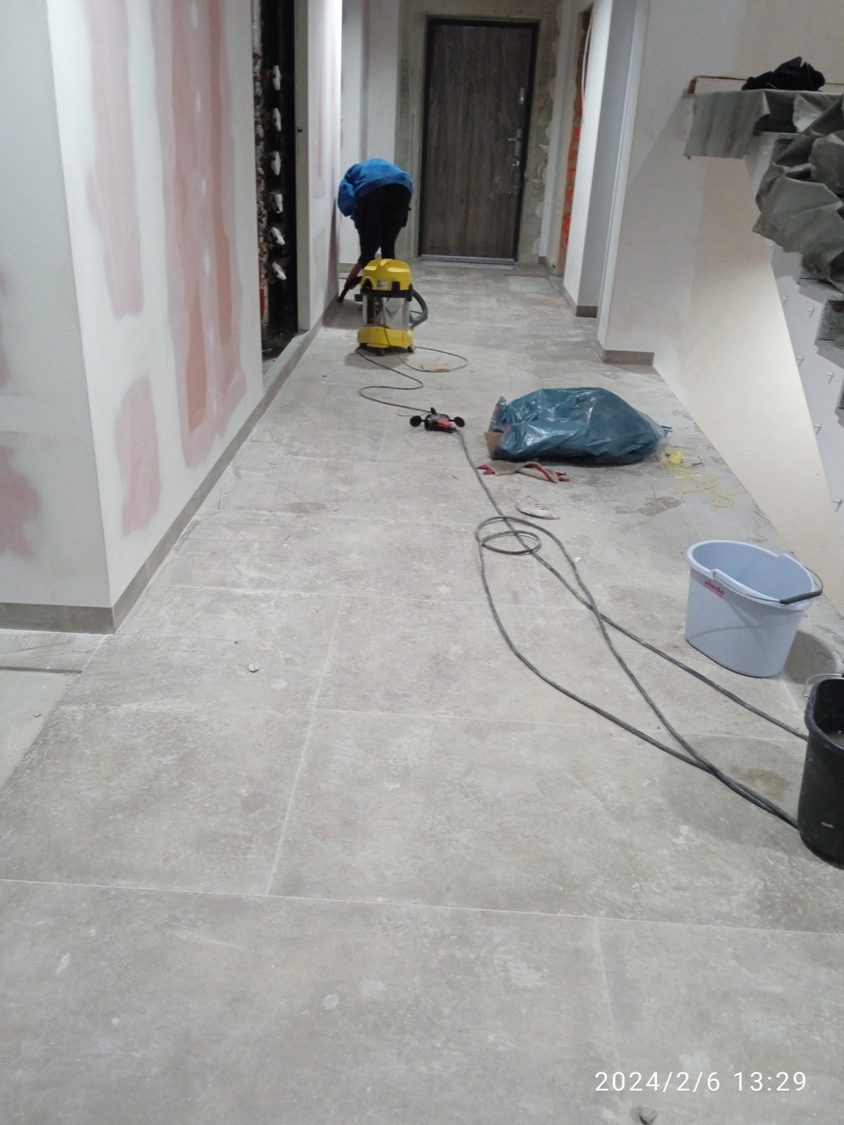 Korytarz w trakcie prac wykończeniowych: ściany częściowo zagruntowane, na podłodze płytki, widoczny odkurzacz przemysłowy, worek na śmieci, wiadro i szlifierka do gipsu.