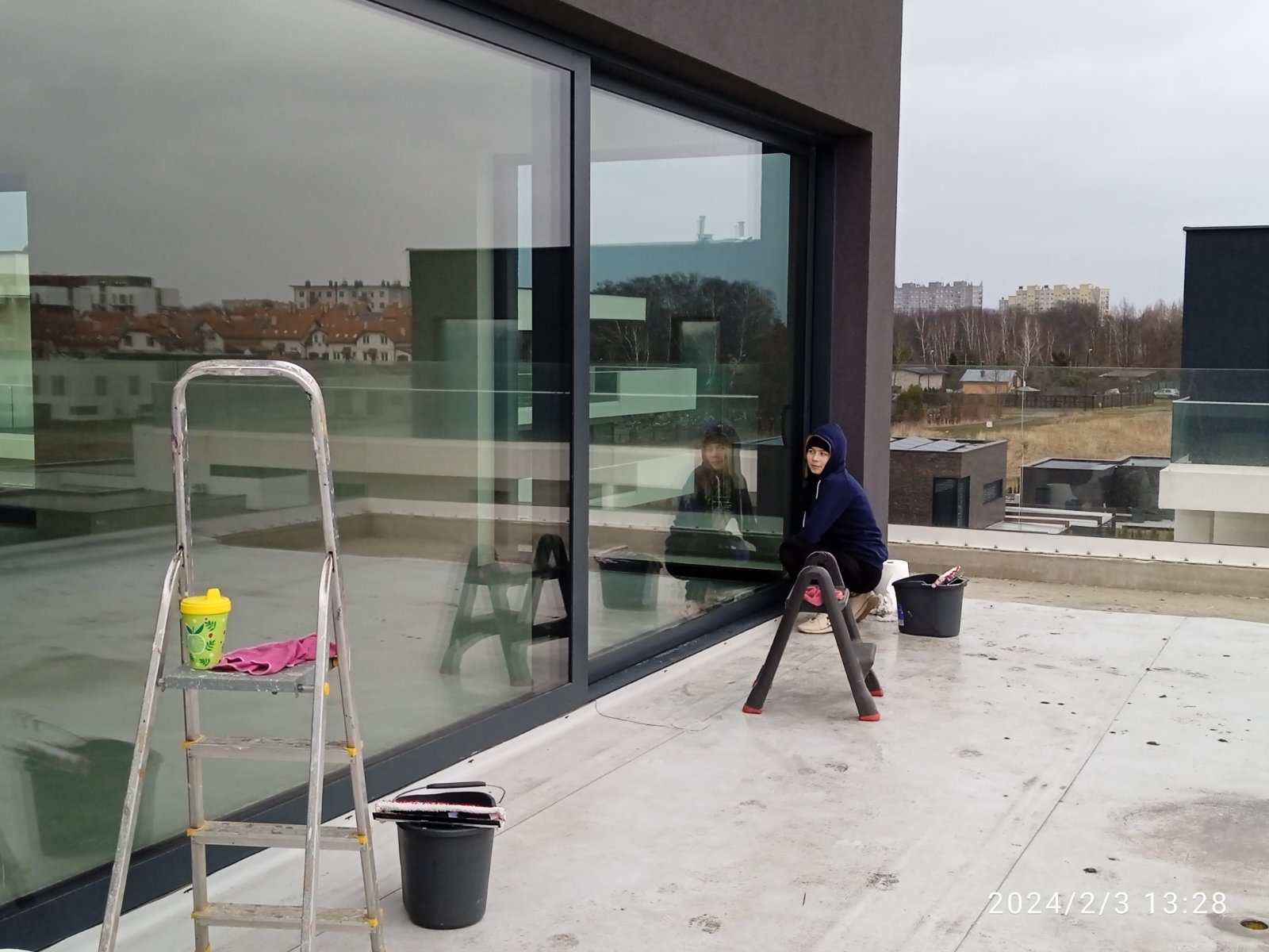 Pracownica w granatowej bluzie z kapturem siedzi na rozkładanej drabince podczas mycia okien w nowoczesnym budynku z dużymi przeszkleniami. Na pierwszym planie drabina z kubkiem i różową ścierką...