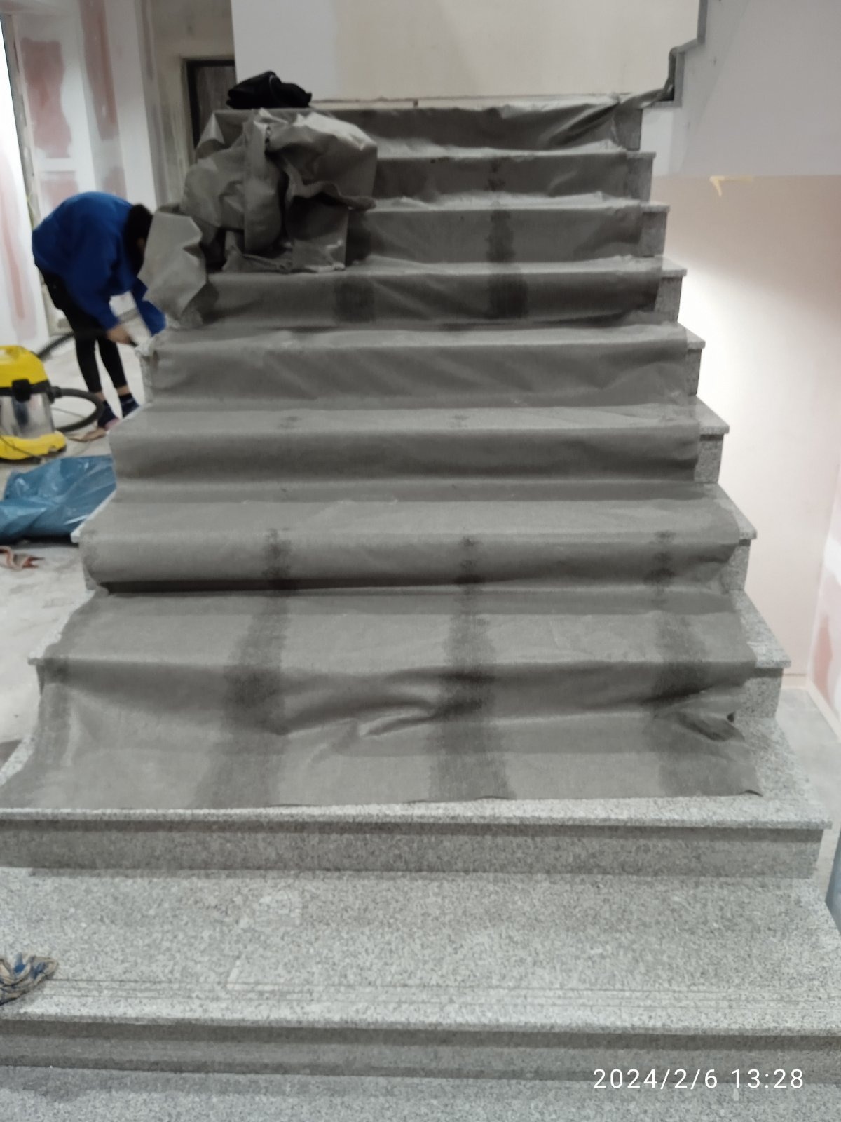 Granitowe schody zabezpieczone folią ochronną podczas prac remontowych, widoczna osoba w niebieskiej bluzie sprzątająca lub przygotowująca do dalszych prac, z odkurzaczem przemysłowym w tle.