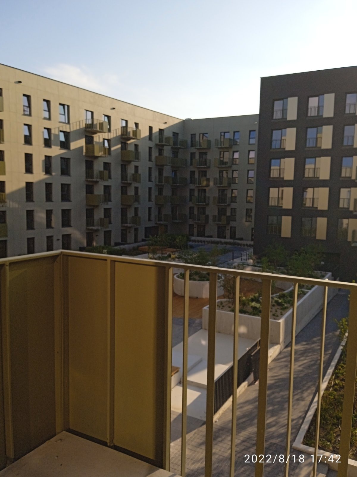 Widok z balkonu na wewnętrzny dziedziniec nowoczesnego kompleksu apartamentów z licznymi balkonami i zielenią w donicach.