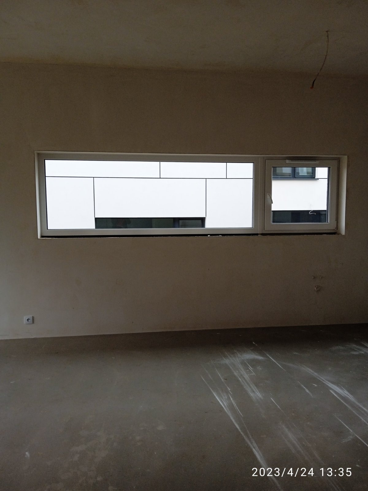 Wnętrze pokoju w stanie surowym z dwoma oknami, widoczne ślady po remoncie na podłodze i ścianach, wystający kabel z sufitu