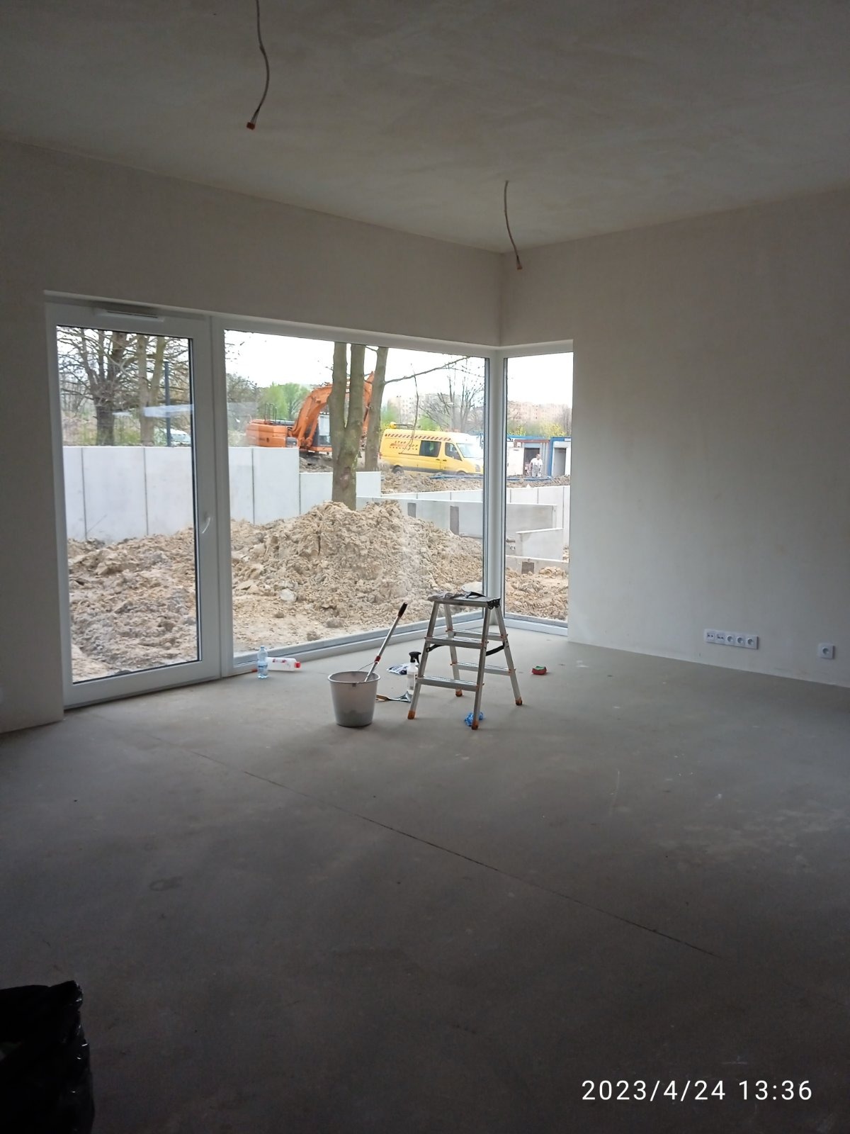 Wnętrze surowego, nieumeblowanego pomieszczenia z dużym oknem, przez które widać plac budowy z koparką i stertą ziemi. Na podłodze stoi wiadro, drabina, mop i butelka z płynem.