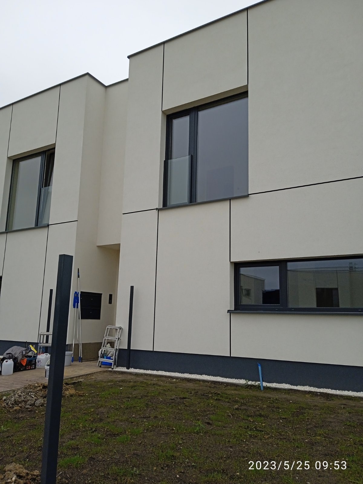 Elewacja nowoczesnego domu z jasnymi panelami i ciemnymi ramami okiennymi, drabiny i narzędzia malarskie w pobliżu, świeżo pomalowany cokół.