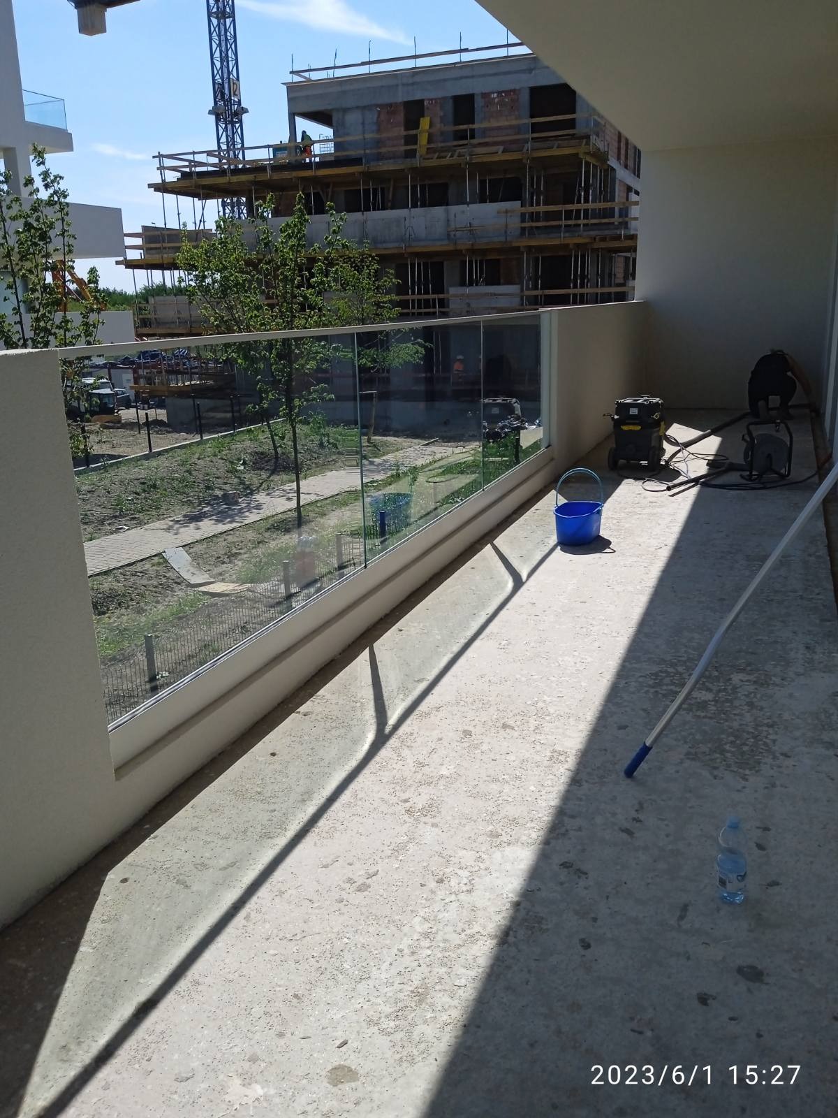 Betonowy balkon z szklaną balustradą w trakcie sprzątania, widoczny odkurzacz przemysłowy, niebieskie wiadro i butelka wody mineralnej, w tle budowa.