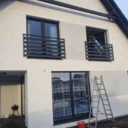 Dwukondygnacyjny dom z szarą stolarką okienną i balkonami z prostymi, poziomymi barierkami; mężczyzna na balkonie w trakcie prac wykończeniowych; drabina oparta o ścianę budynku.