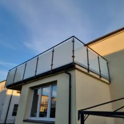 Szklana balustrada balkonowa z czarnymi profilami, widok z dołu na tle błękitnego nieba, odbicie budynku w oknach.