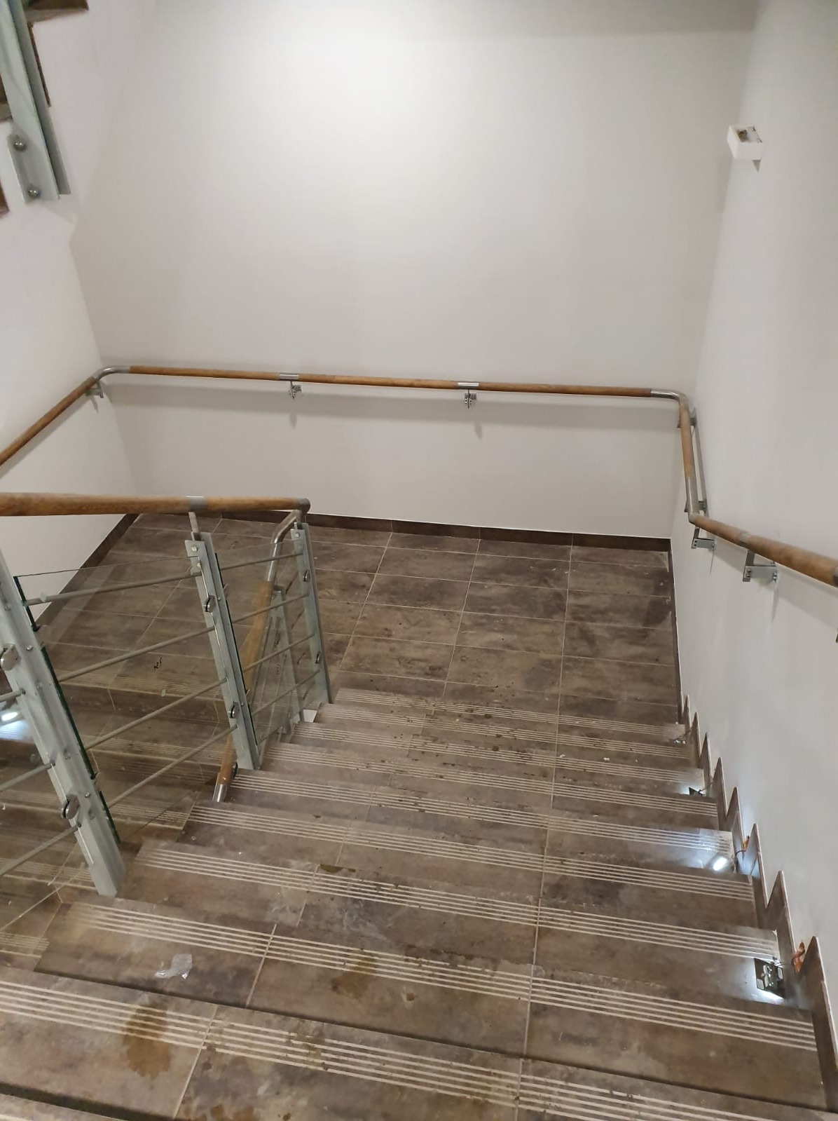 Wewnętrzne schody z brązowymi płytkami i balustradą ze szkła oraz drewnianą poręczą przymocowaną do ściany za pomocą metalowych uchwytów.