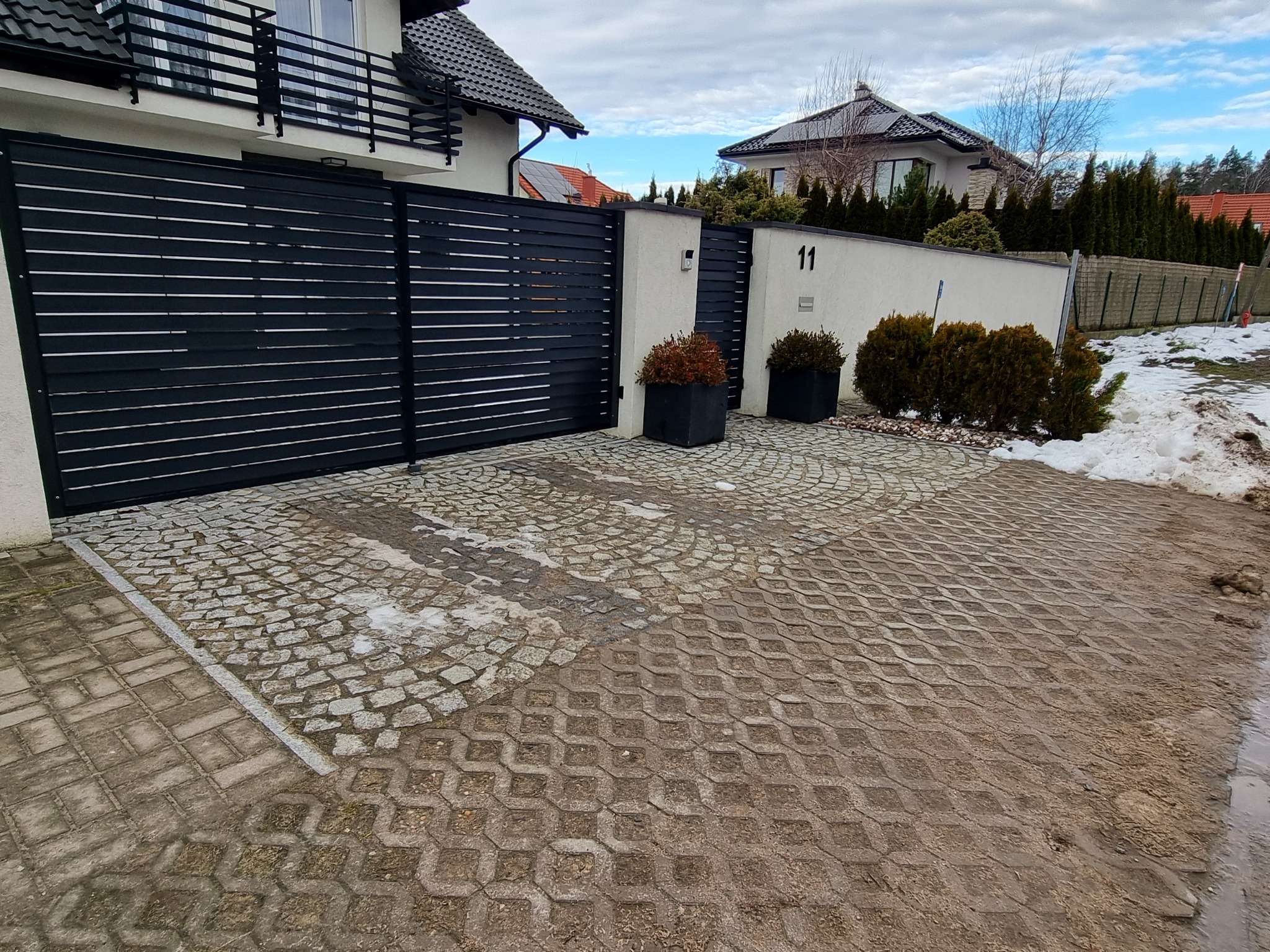 Nowoczesna, pozioma brama wjazdowa z czarnych, płaskich listew, z numerem domu '11' na białym murze, otoczona dekoracyjnymi krzewami w czarnych donicach i brukowaną nawierzchnią z kostki.