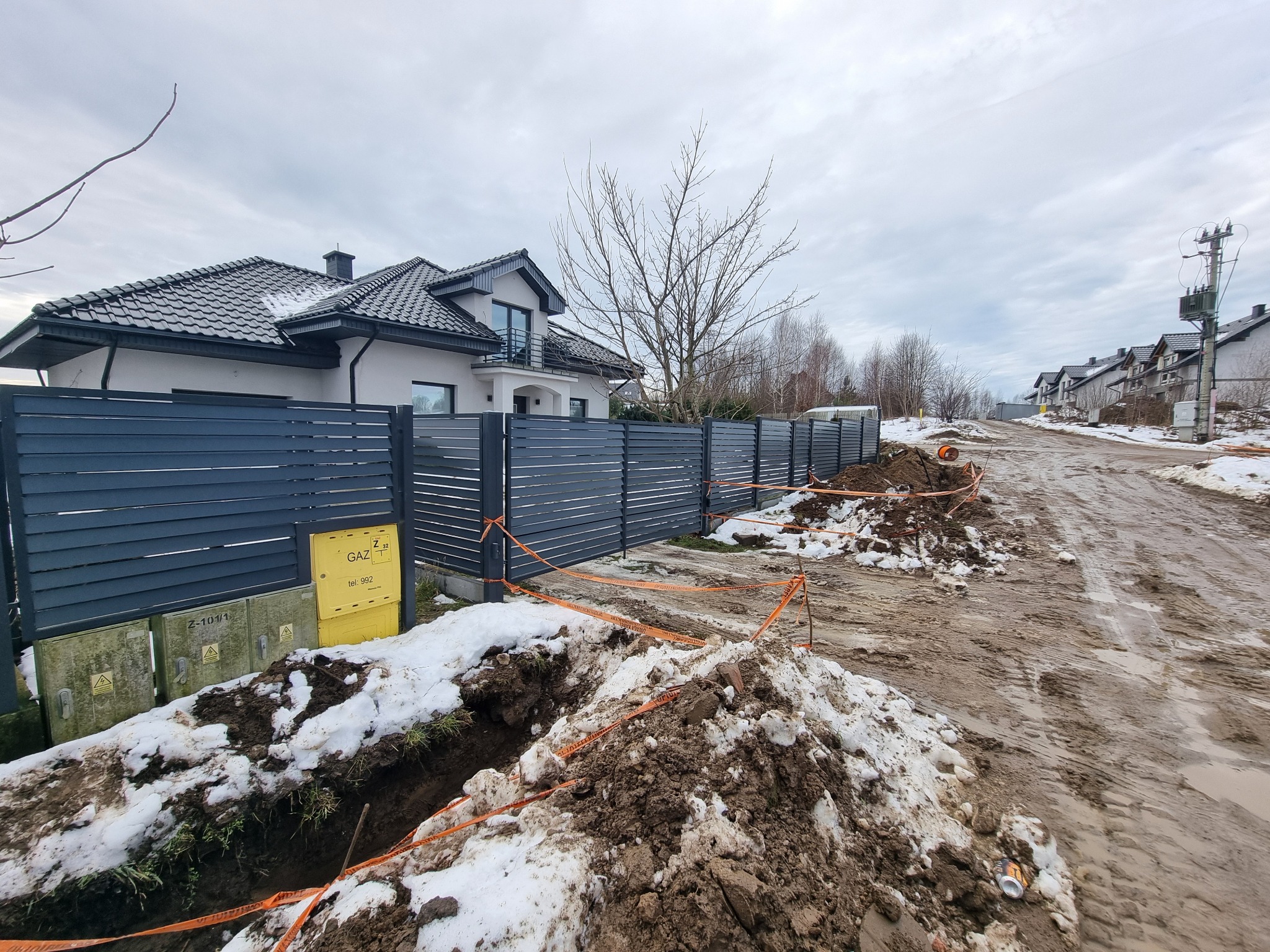 Szare ogrodzenie panelowe z poziomych listew, widoczne skrzynki gazowe i rozkopana ziemia z resztkami śniegu sugerująca prace ziemne w toku.
