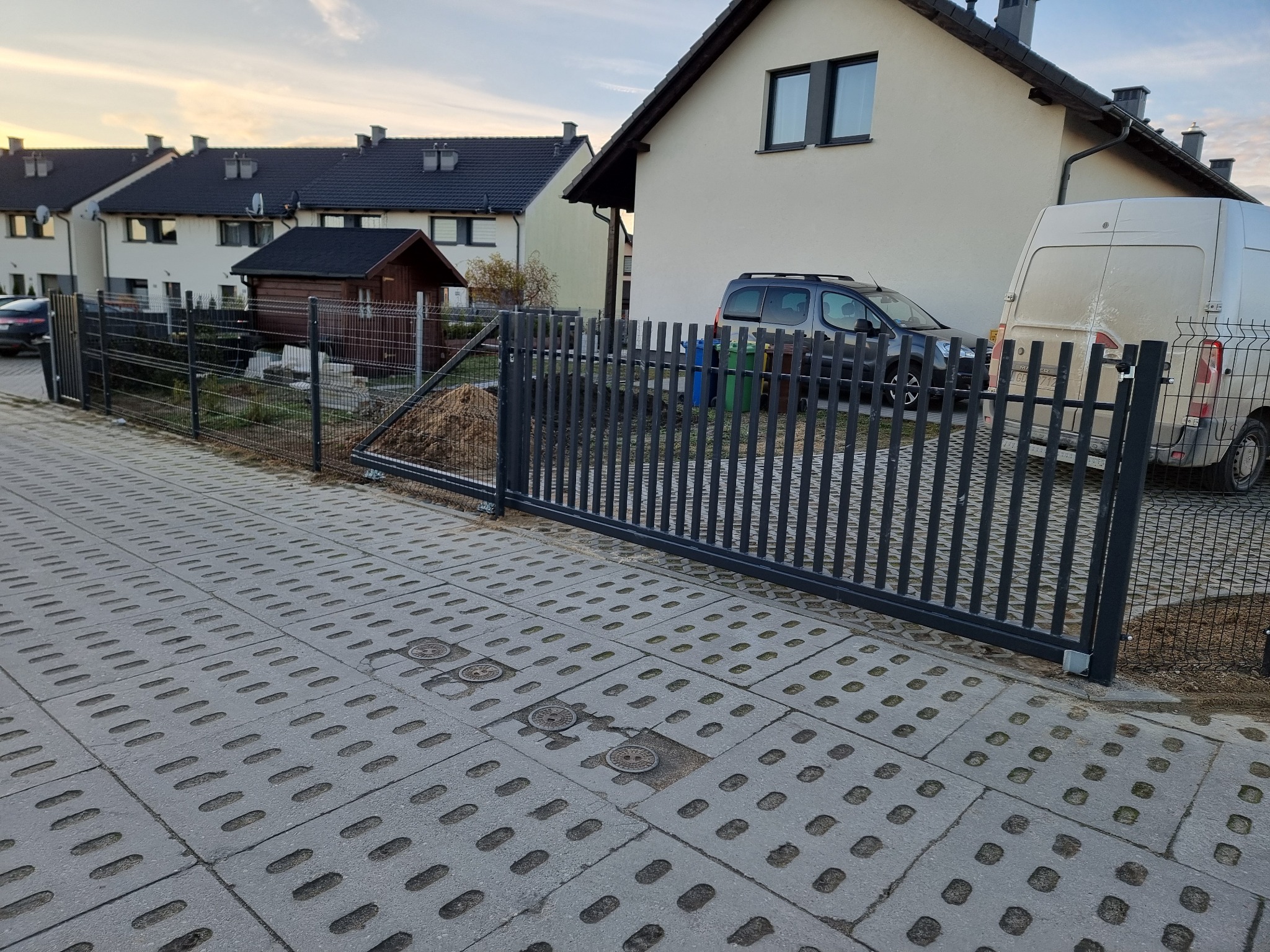Nowoczesna, przesuwna brama wjazdowa z pionowych profili, widoczna na tle domu jednorodzinnego z podjazdem wyłożonym ażurową kostką brukową.