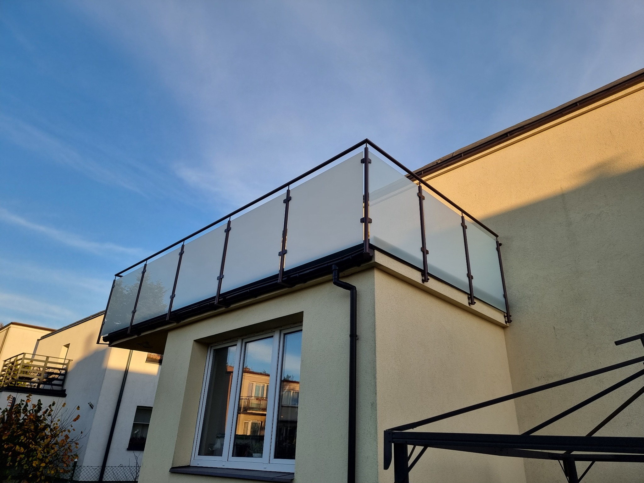 Szklana balustrada balkonowa z czarnymi profilami, widok z dołu na tle błękitnego nieba, odbicie budynku w oknach.