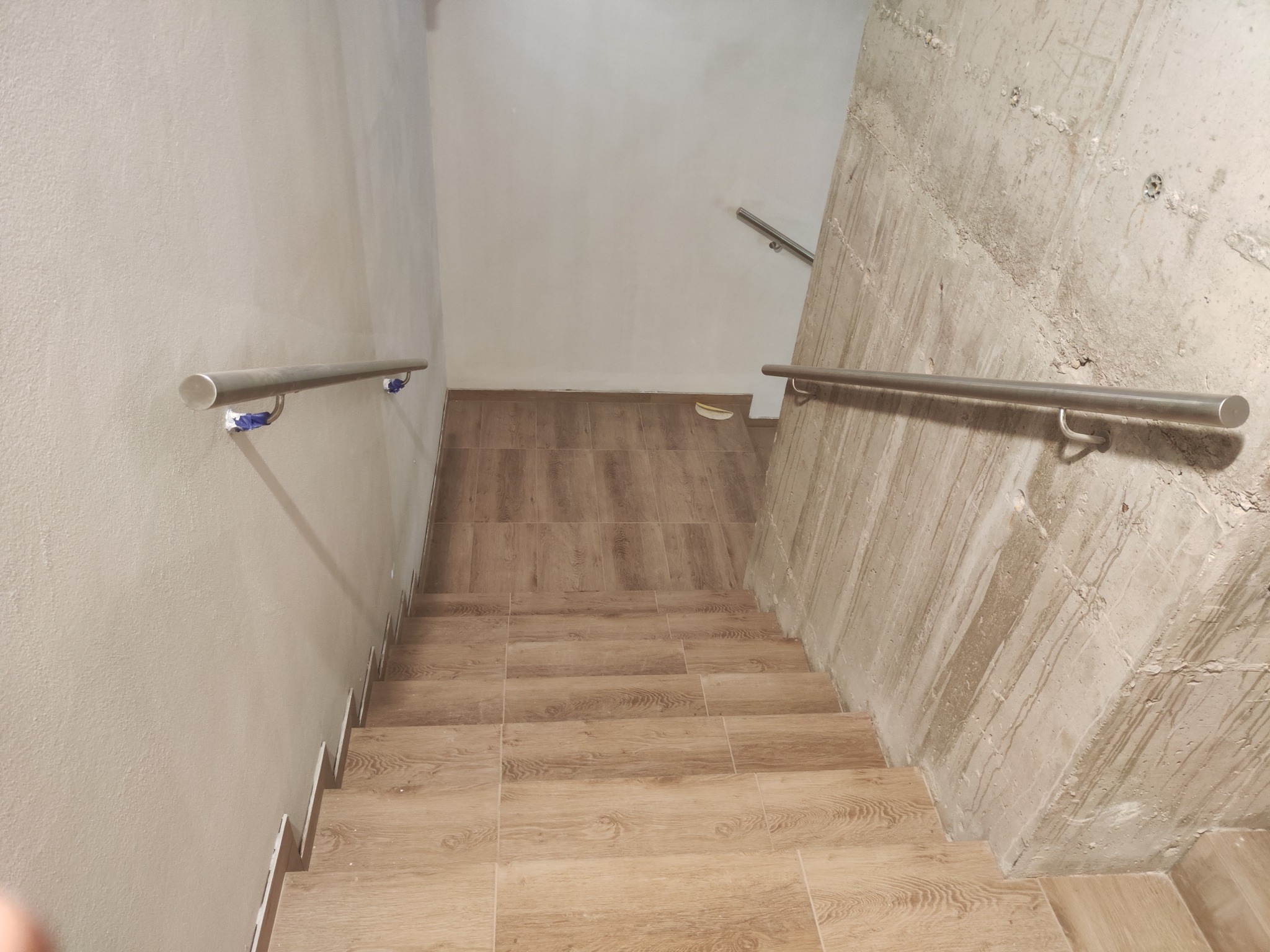 Klatka schodowa w surowym stanie z wykończonymi drewnopodobnymi płytkami stopniami i zamontowaną metalową poręczą na ścianie z betonu architektonicznego oraz na ścianie pomalowanej na jasny kolor...