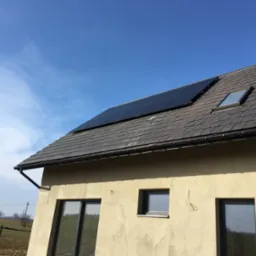 Panele fotowoltaiczne na dachu domu z szarej dach&oacute;wki i oknem dachowym, widok z dołu na jasną elewację i okna.