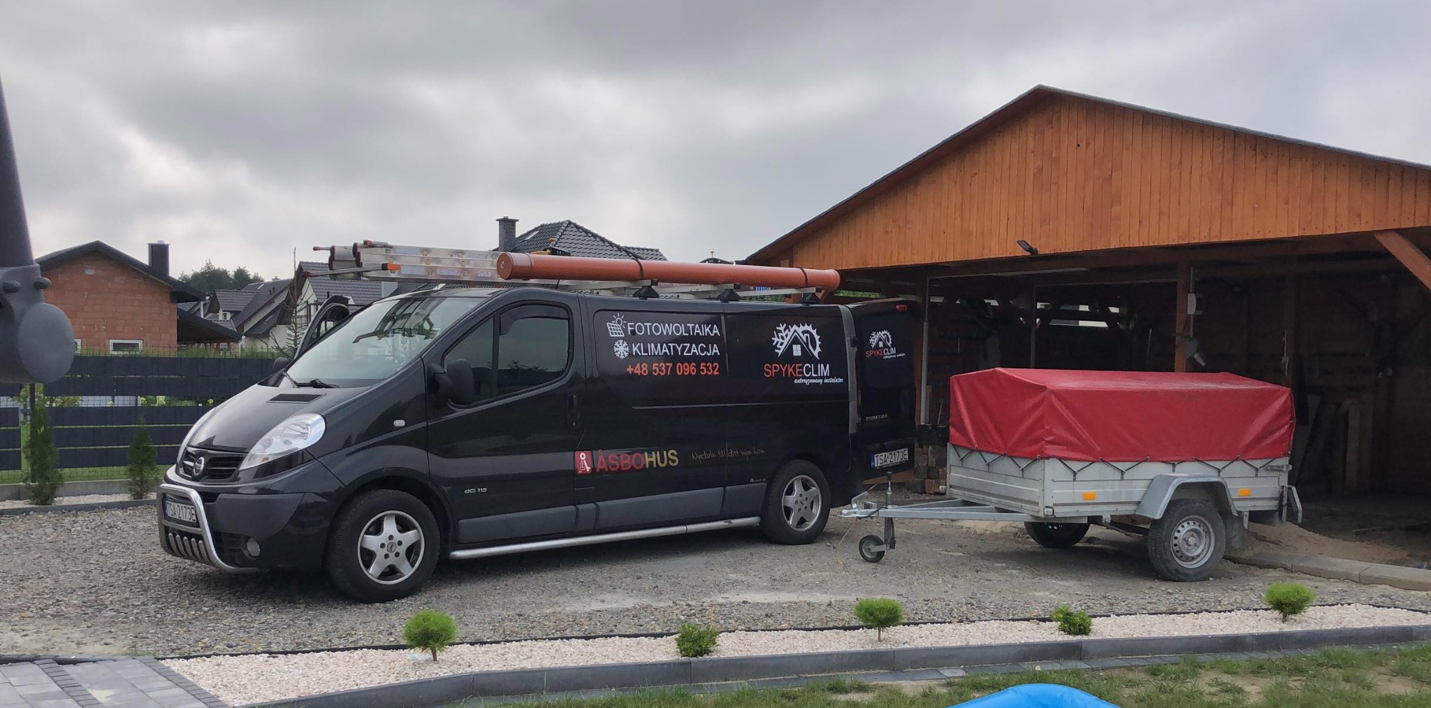 Czarny van z drabiną na dachu i przyczepką, z logo firmy oferującej usługi fotowoltaiki i klimatyzacji, zaparkowany na żwirowym podjeździe przed drewnianą wiatą.