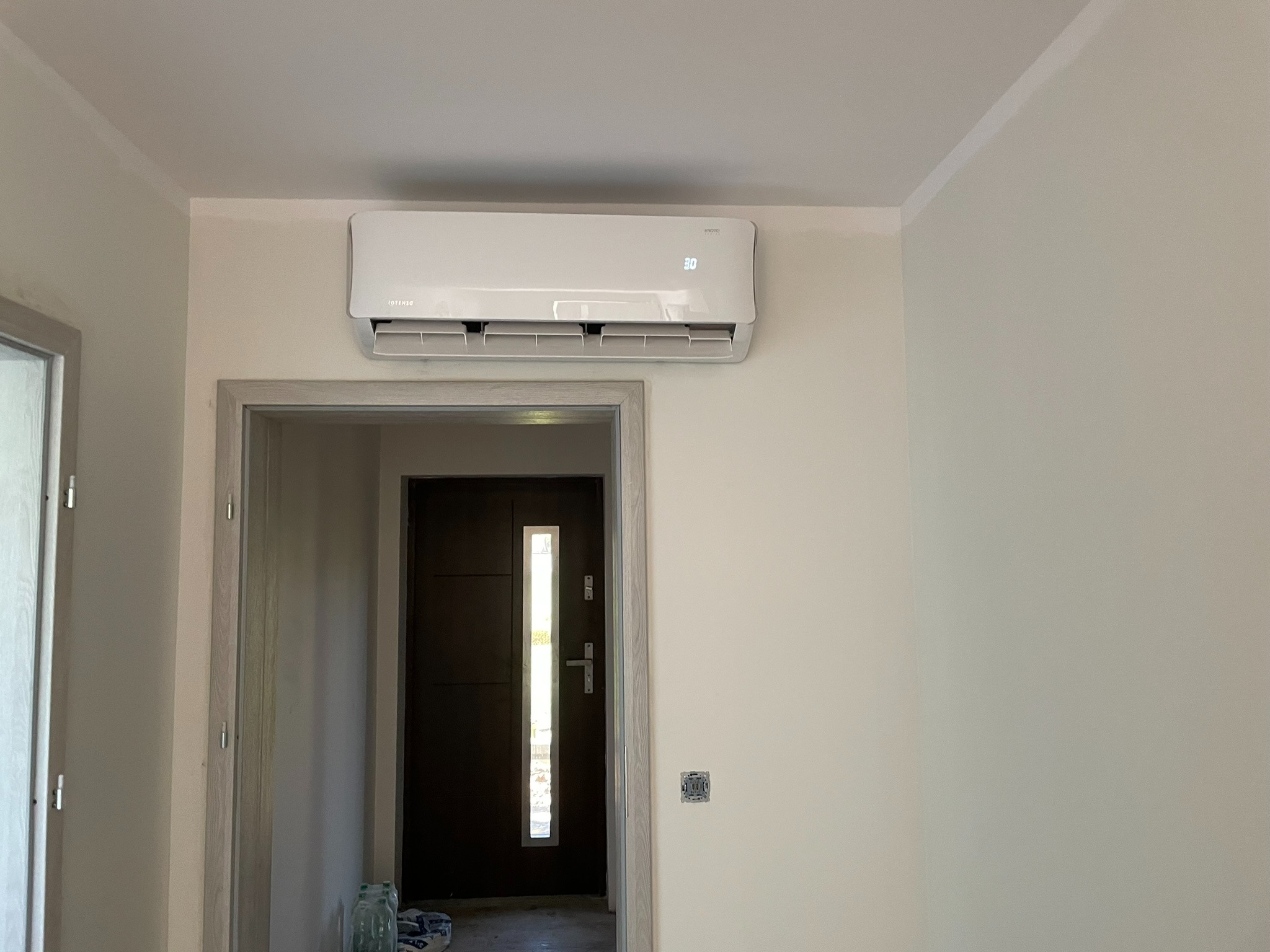Biały klimatyzator ścienny Rotenso zamontowany nad ościeżnicą drzwi wejściowych w jasnym pomieszczeniu, widoczny wyświetlacz temperatury 30 stopni.