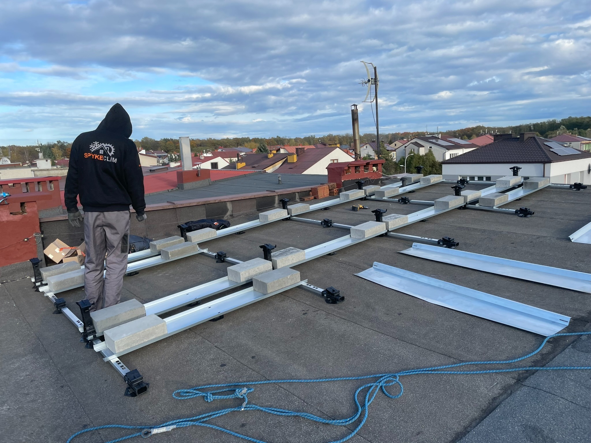 Montaż konstrukcji wsporczej pod panele fotowoltaiczne na dachu budynku; pracownik w bluzie z logo firmy na tle zabudowy jednorodzinnej.