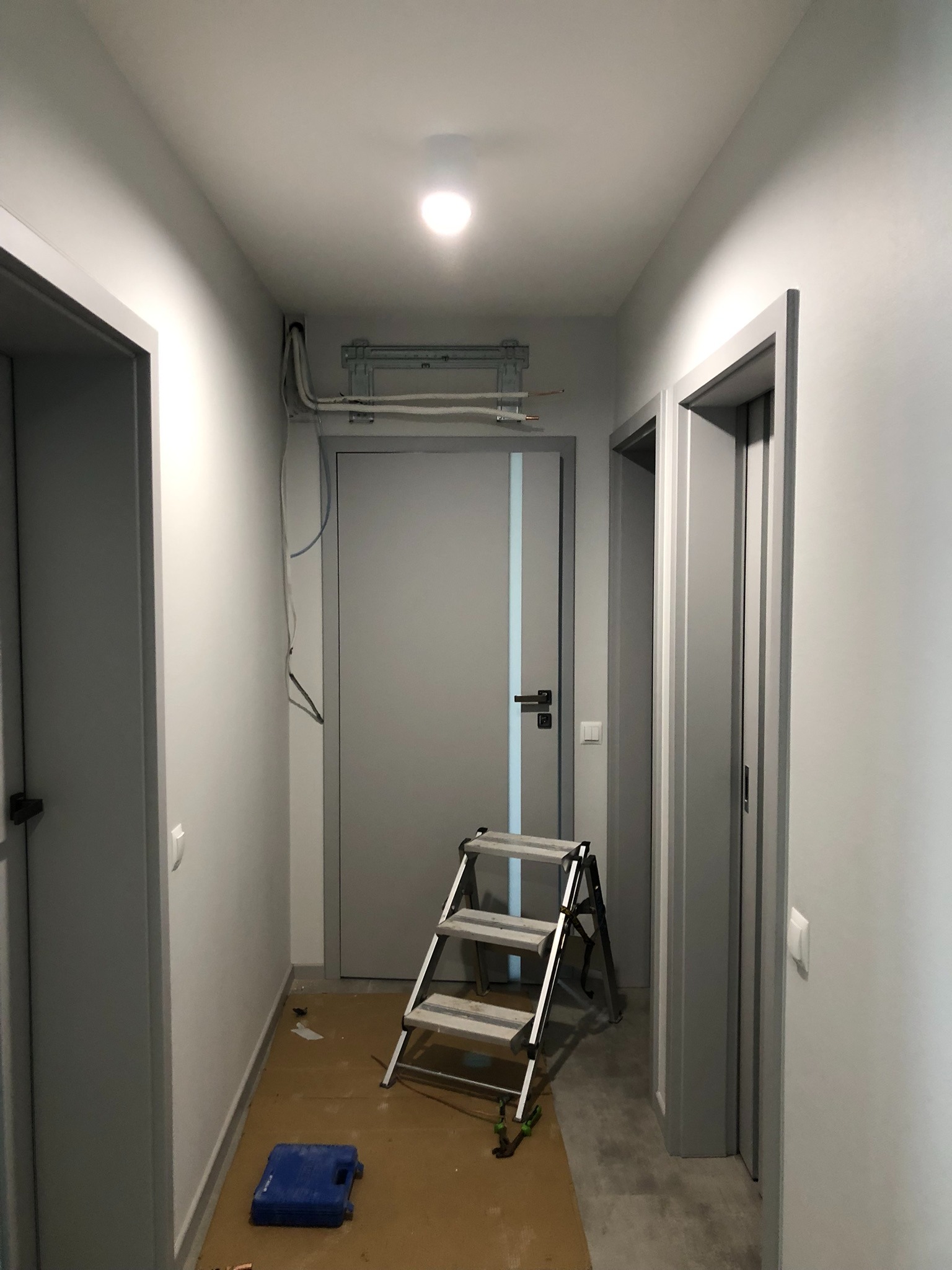 Korytarz w trakcie montażu klimatyzacji: widoczny wspornik na ścianie pod sufitem, przewody, drabina aluminiowa, niebieska skrzynka narzędziowa i zabezpieczona podłoga.
