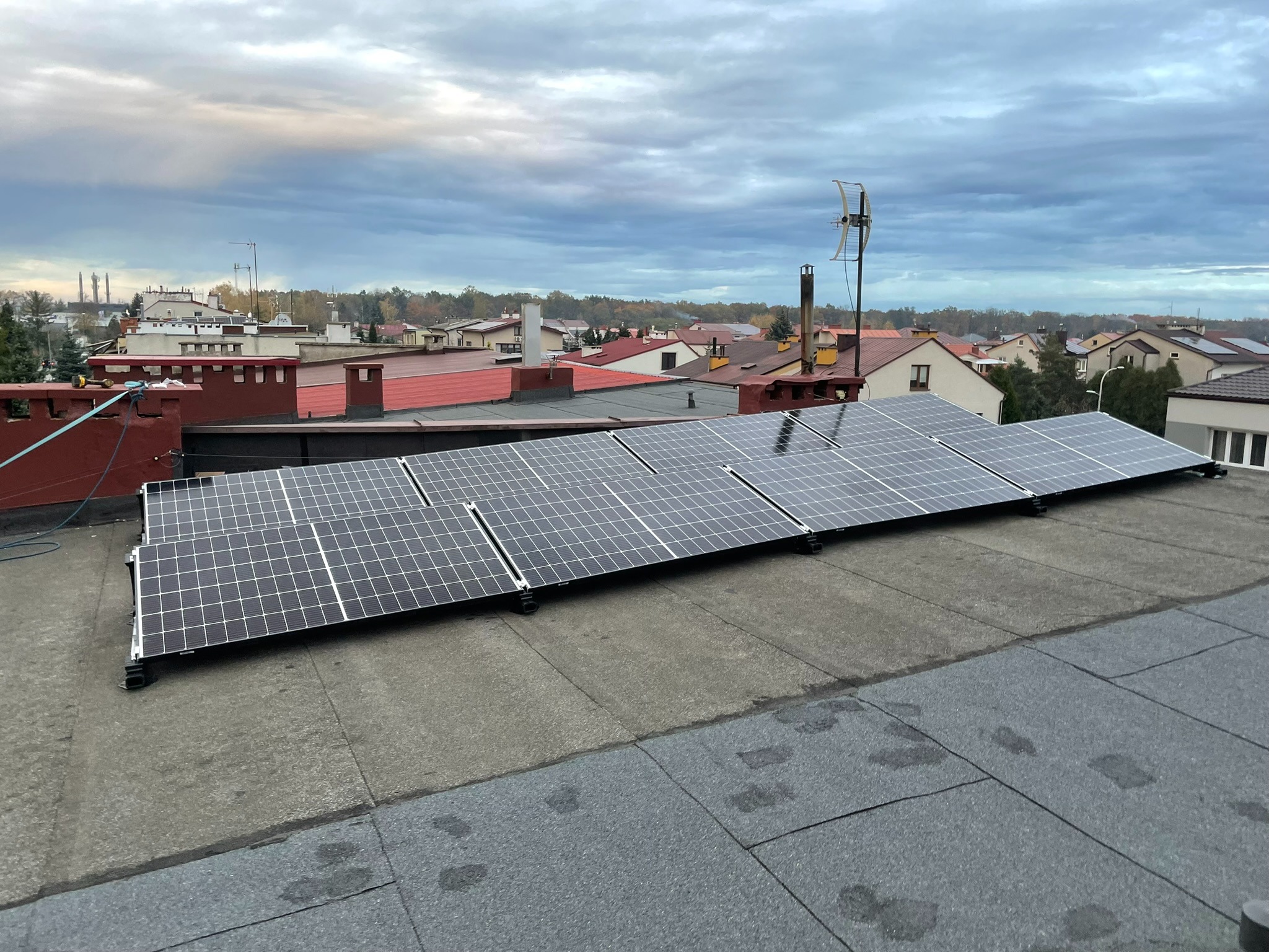 Panele fotowoltaiczne zamontowane na płaskim dachu budynku z widokiem na okoliczną zabudowę i pochmurne niebo.