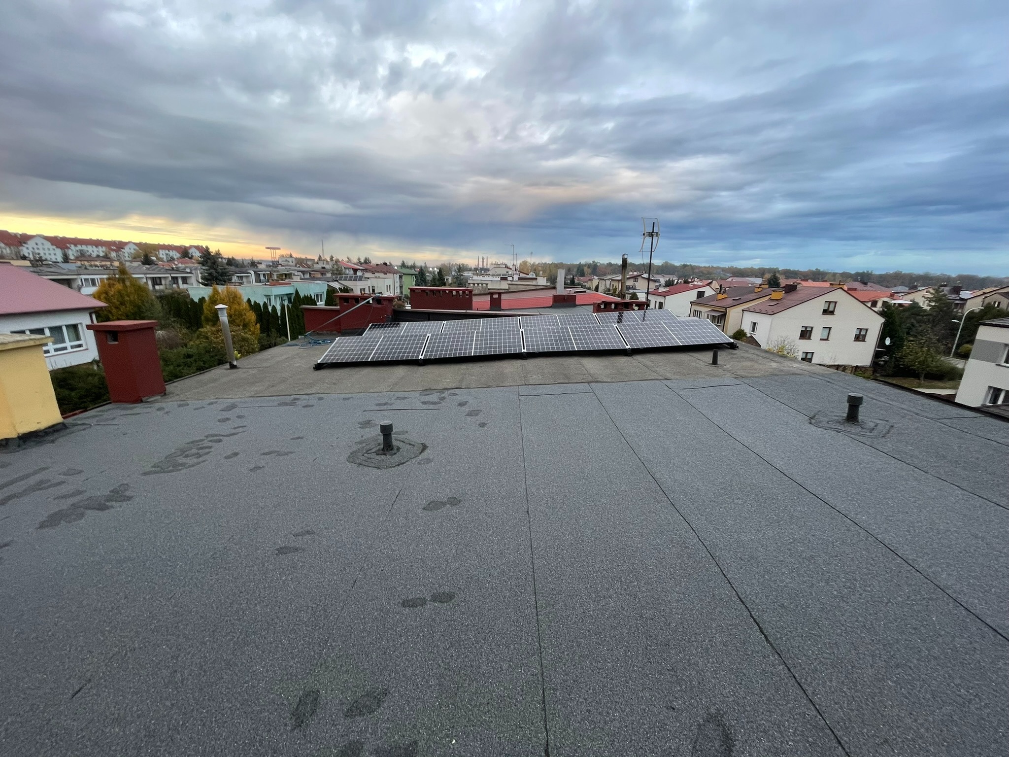 Panel fotowoltaiczny na płaskim dachu budynku w zabudowie miejskiej, widok z góry na tle pochmurnego nieba.