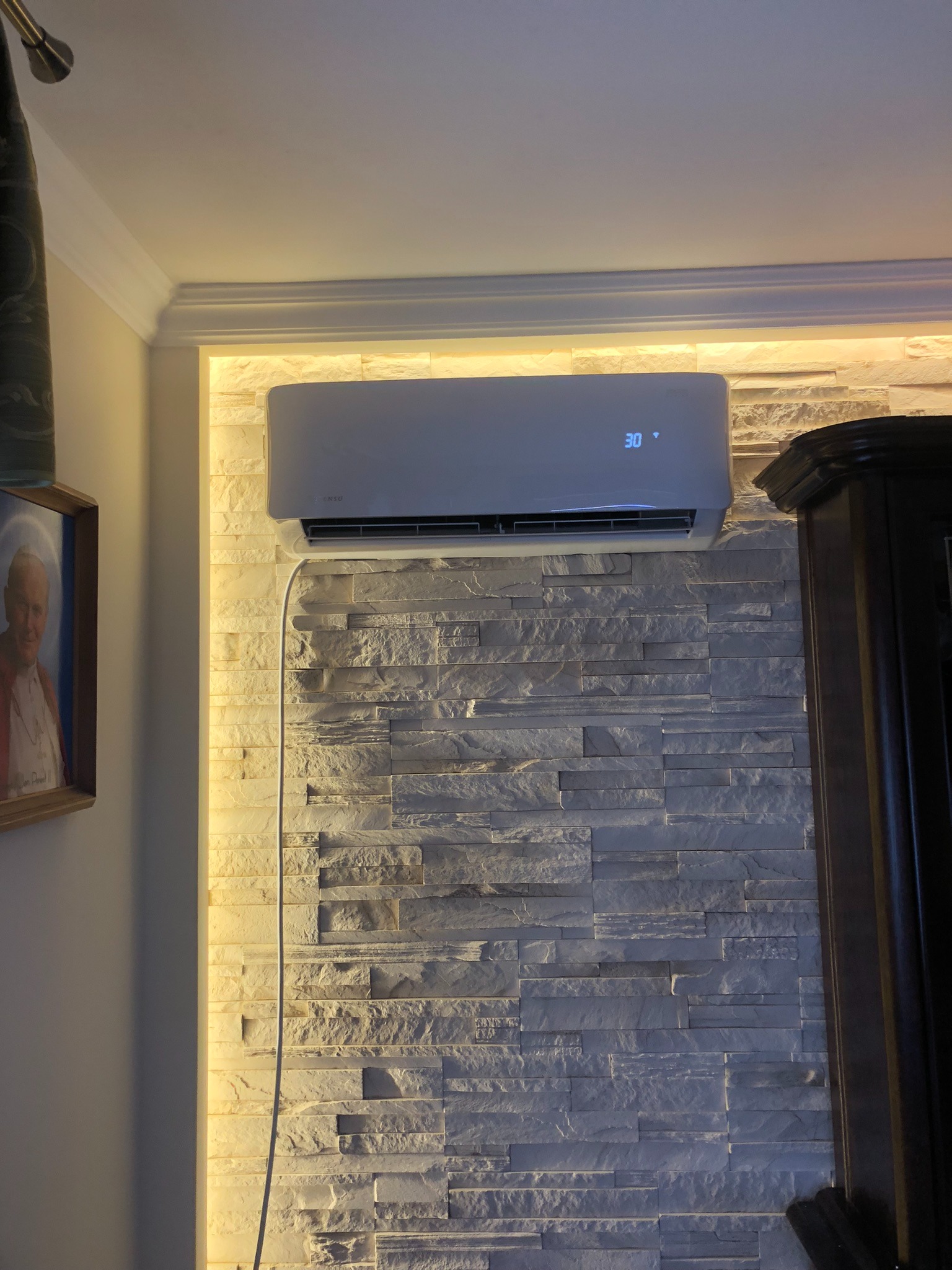 Biały klimatyzator ścienny z wyświetlaczem ustawionym na 30 stopni, zamontowany na ścianie z dekoracyjnej szarej cegły, podświetlonej od góry ciepłym światłem LED.
