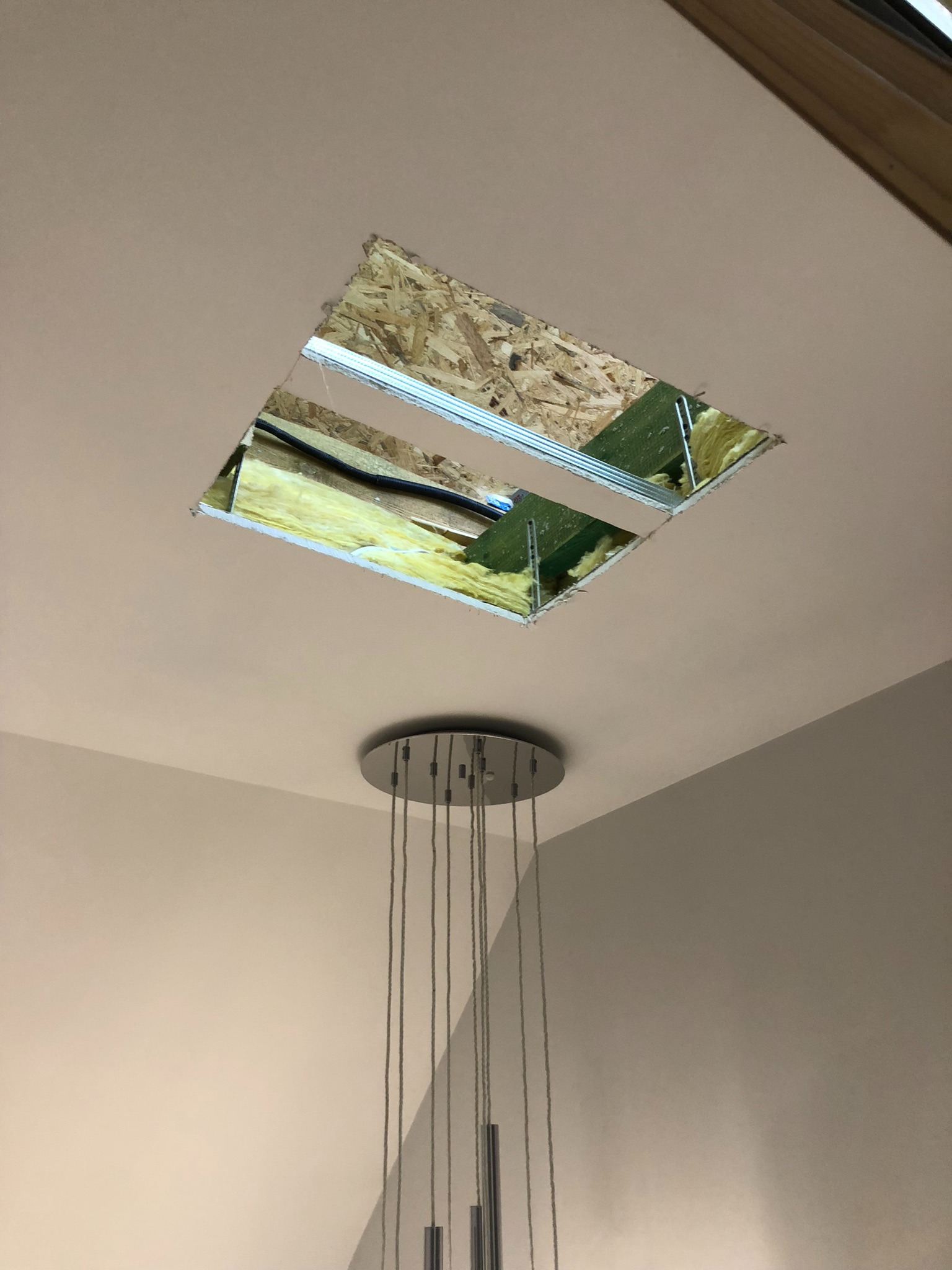 Otwór w suficie odsłaniający izolację z wełny mineralnej, widoczne elementy konstrukcji drewnianej i instalacji elektrycznej, poniżej nowoczesna lampa wisząca.