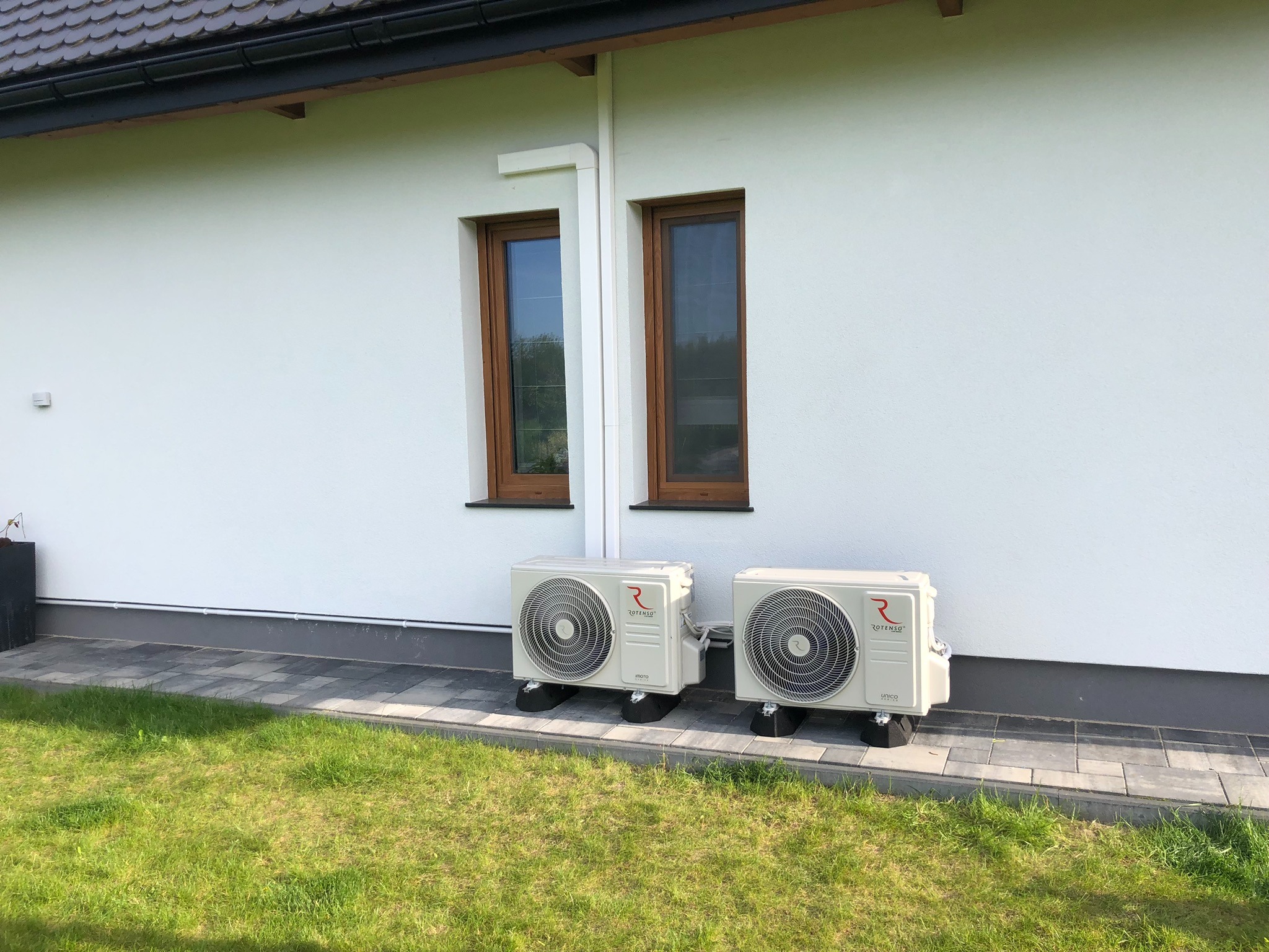 Dwa zewnętrzne agregaty klimatyzacji Rotenso zamontowane na niskich podstawach przy białej ścianie domu, widoczne dwa brązowe okna, zielony trawnik.