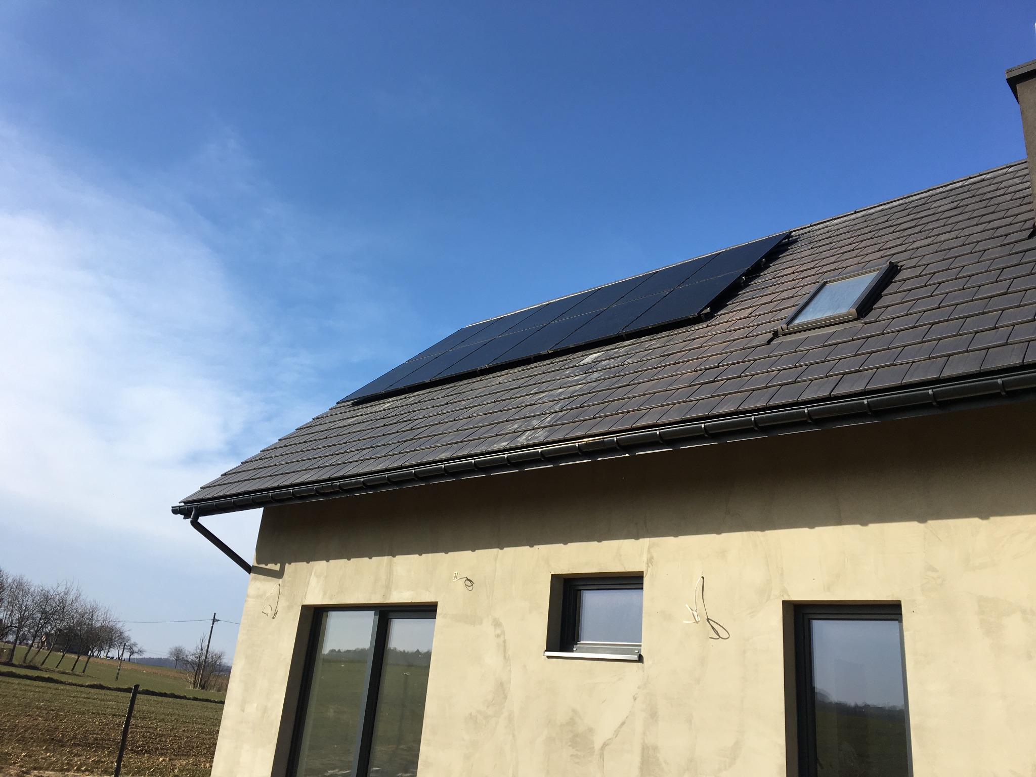 Panele fotowoltaiczne na dachu domu z szarej dachówki i oknem dachowym, widok z dołu na jasną elewację i okna.