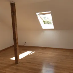 Poddasze z drewnianą podłogą, belką konstrukcyjną i oknem dachowym wpuszczającym światło. Widoczne gniazdko elektryczne na ścianie.