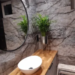 Nowoczesna łazienka z umywalką nablatową, drewnianym blatem, marmurowymi ścianami i okrągłym lustrem z czarną ramą. W tle widoczna roślina w srebrnej doniczce i drabinka.