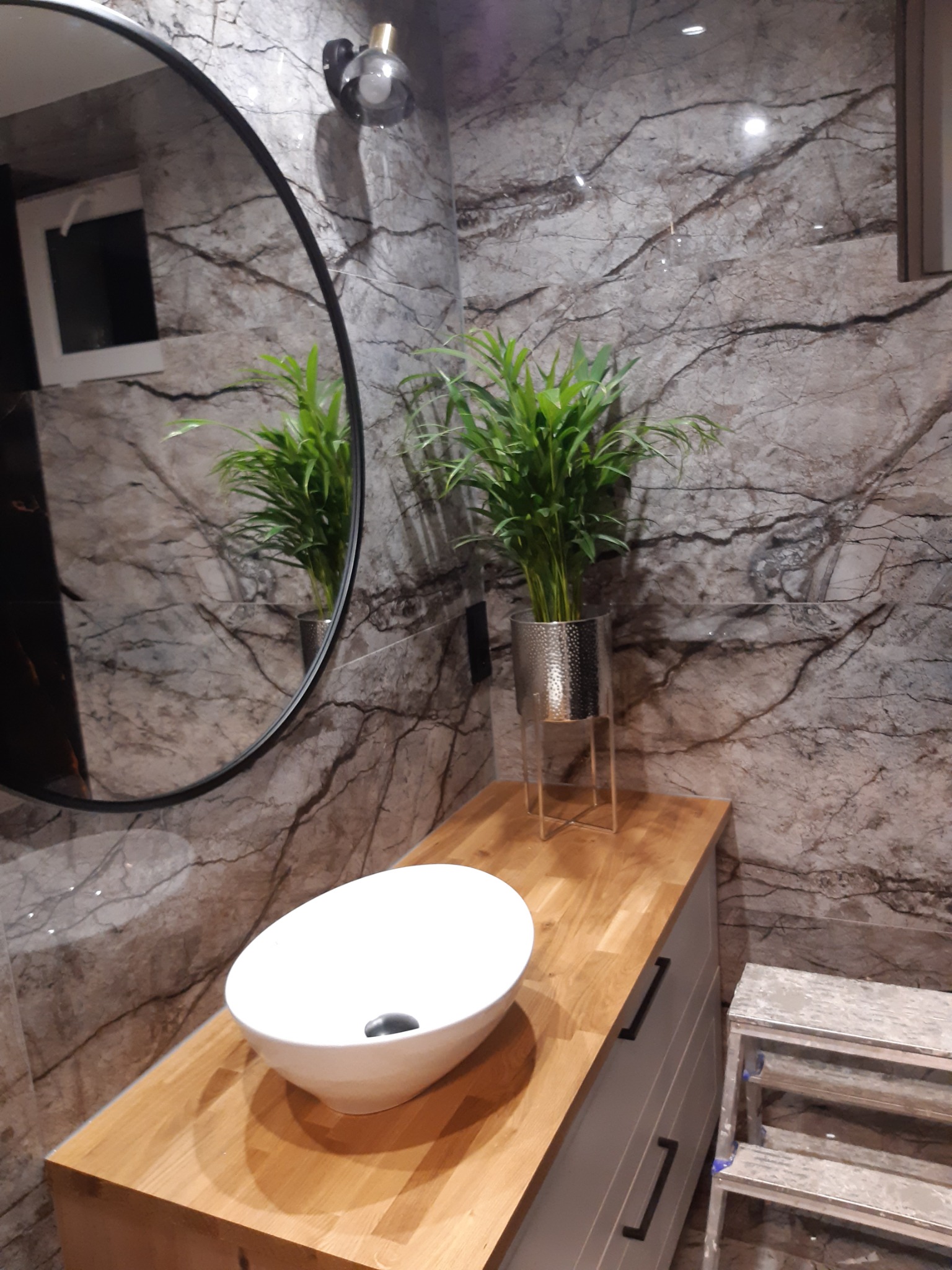 Nowoczesna łazienka z marmurowymi ścianami, okrągłym lustrem, białą umywalką nablatową, drewnianym blatem, szafką z szufladami i rośliną w srebrnej doniczce.