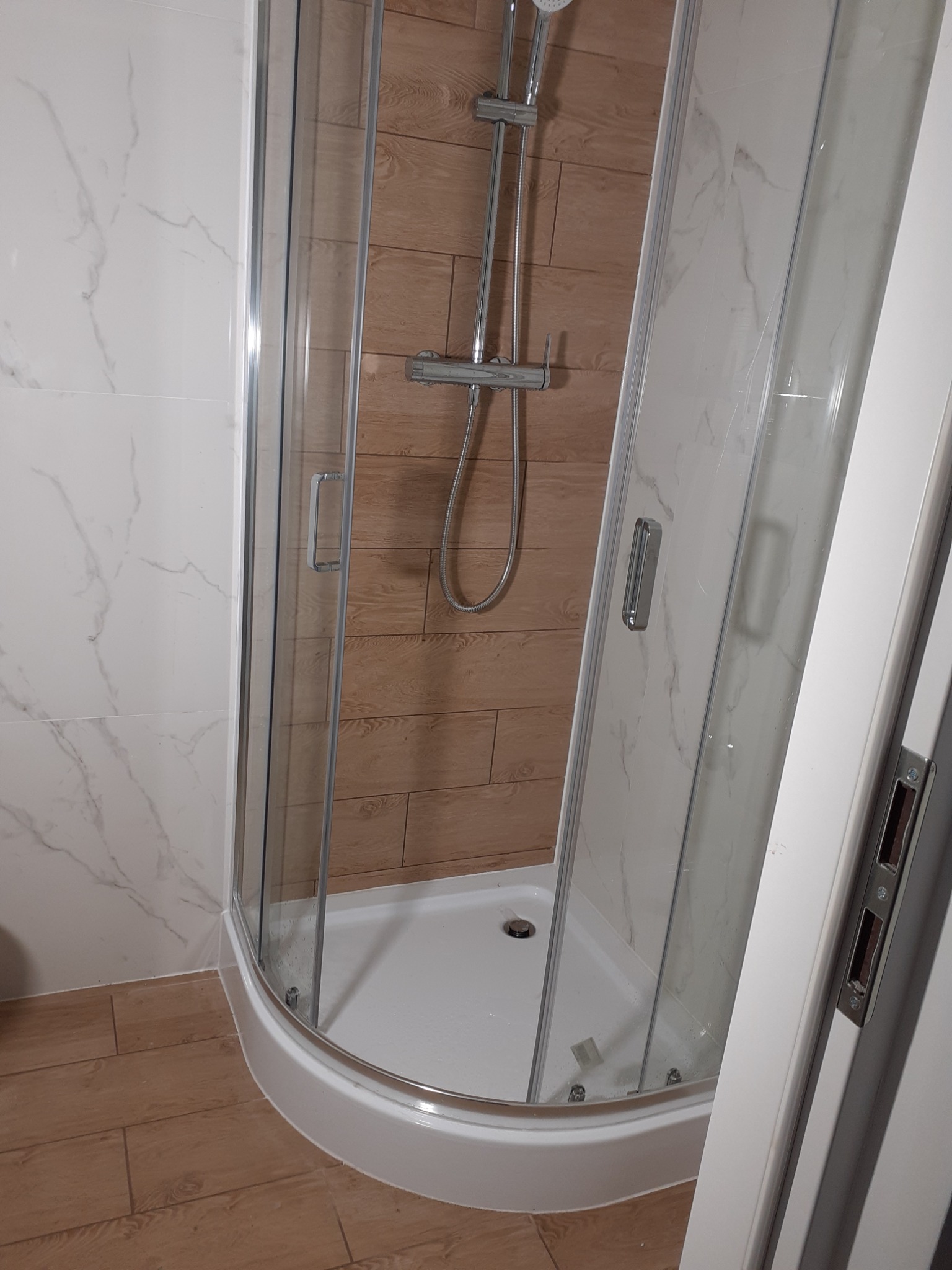 Kabina prysznicowa z przesuwnymi drzwiami, wykończona płytkami imitującymi drewno na jednej ze ścian i marmur na pozostałych, widok zza uchylonych drzwi.