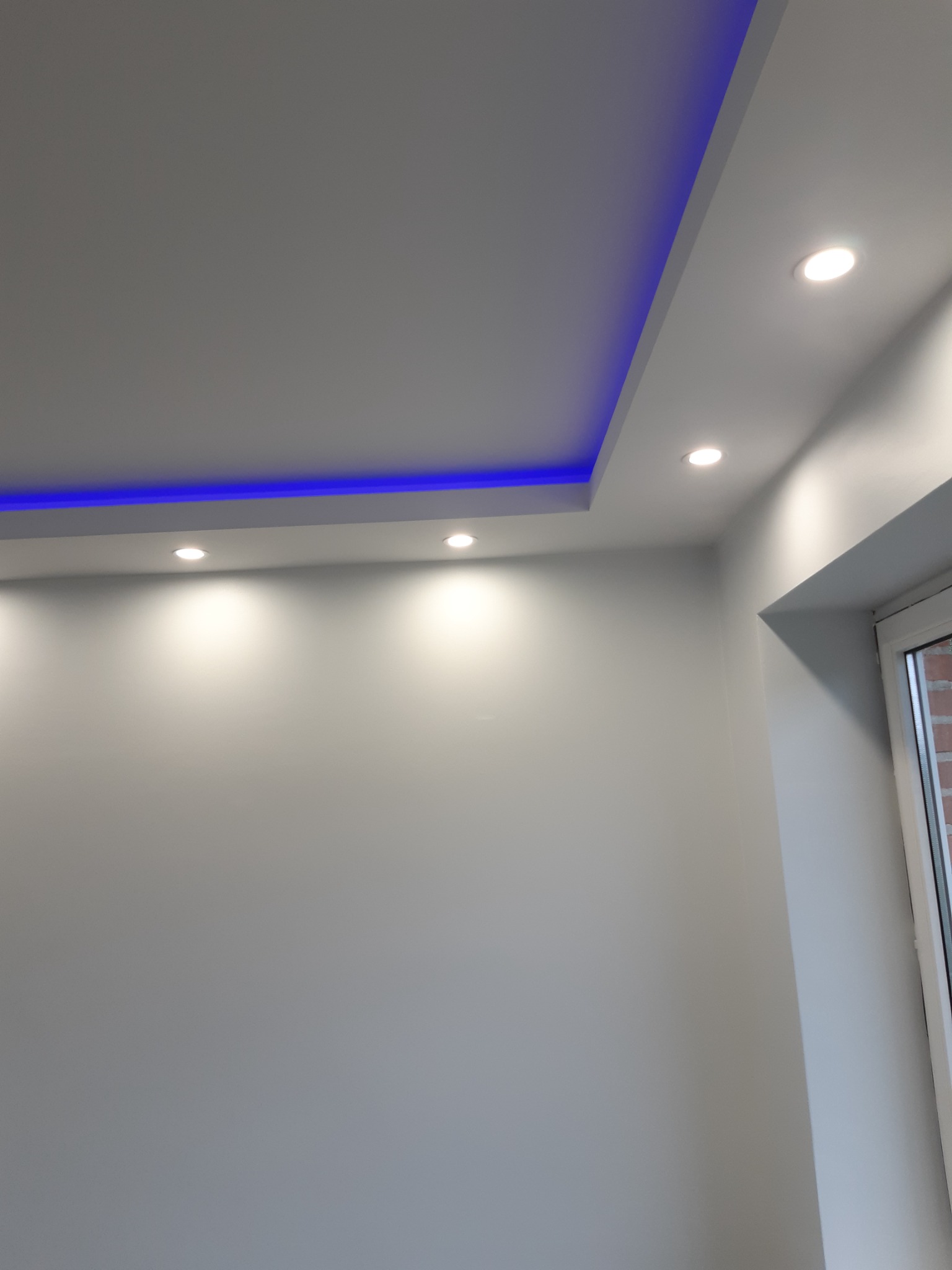 Szare wnętrze z podwieszonym sufitem, oświetleniem LED w kolorze niebieskim i trzema punktowymi lampami halogenowymi, widoczny fragment okna.