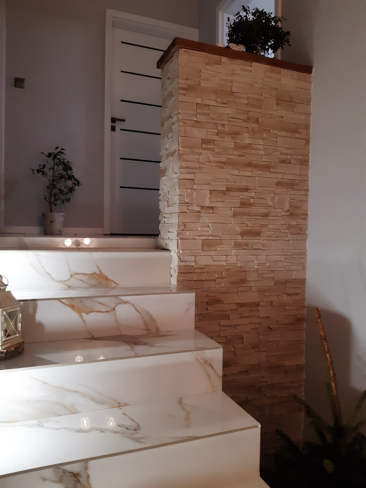 Schody wyłożone białymi płytkami imitującymi marmur, z oświetleniem punktowym, obok ściana z dekoracyjnej cegły w odcieniach beżu i brązu oraz białe drzwi z czarnymi akcentami.