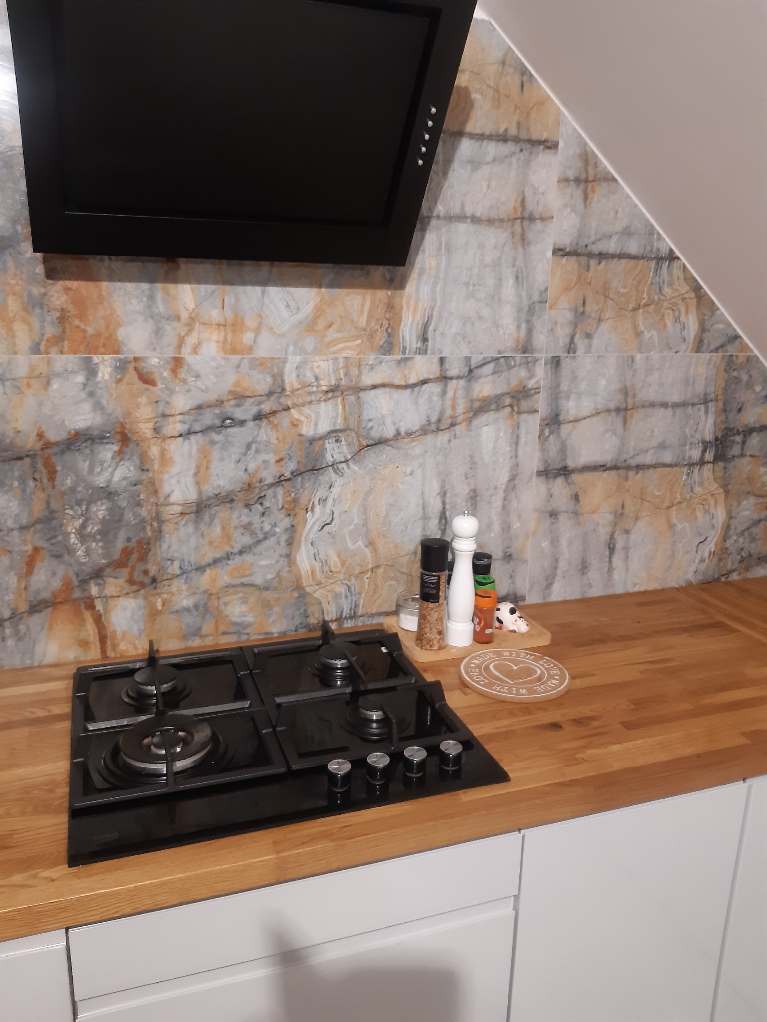 Nowoczesna kuchnia z czarną płytą gazową, drewnianym blatem, marmurowymi płytkami na ścianie i czarnym okapem; przyprawy i dekoracje na blacie.