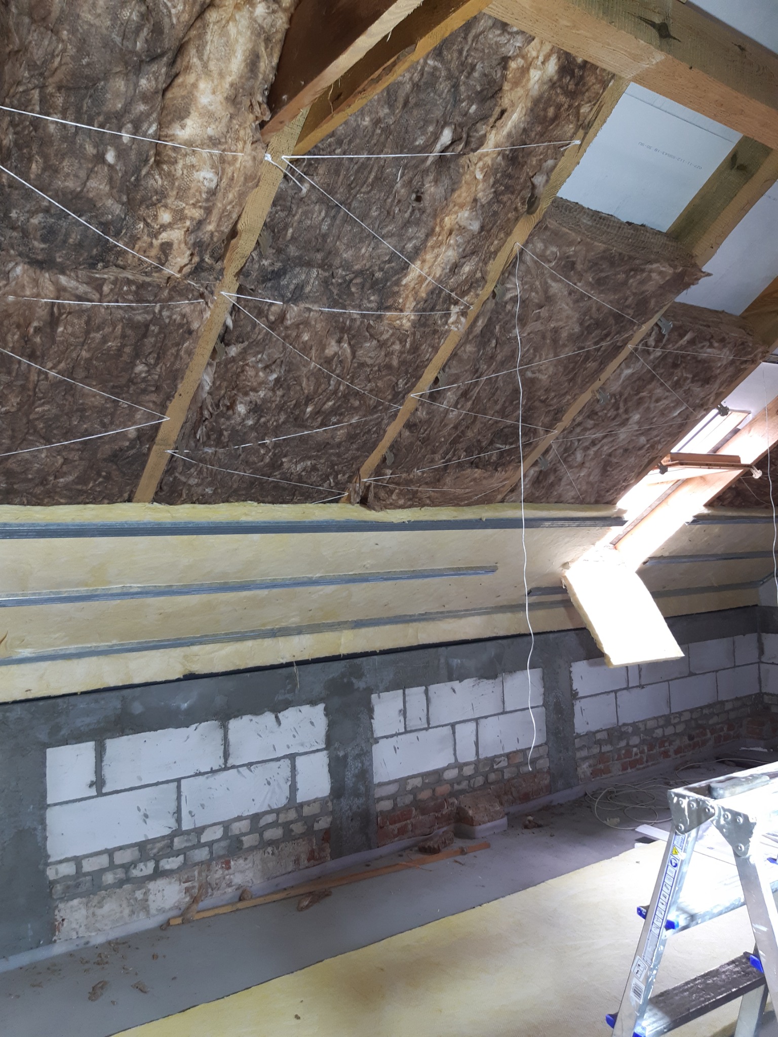 Poddasze w trakcie izolacji termicznej: wełna mineralna między krokwiami, zamocowana sznurkami, ściany z bloczków betonowych i cegieł w surowym stanie, widoczna drabina.