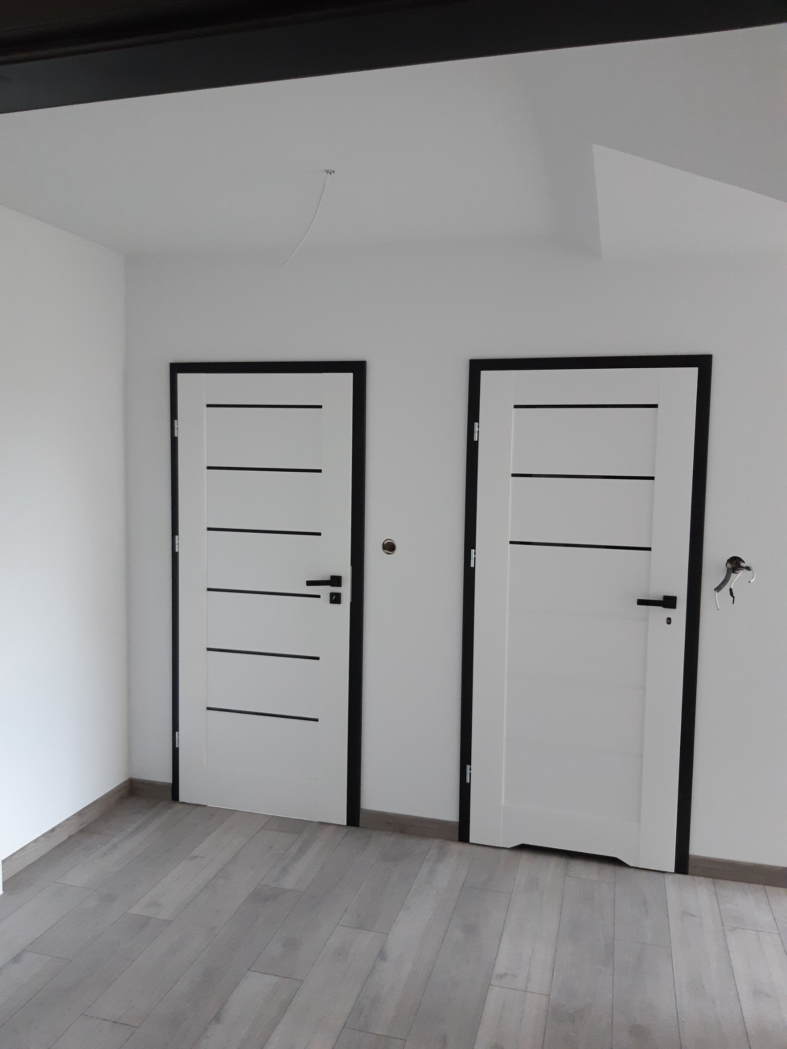 Dwa białe drzwi z czarnymi ramami i poziomymi paskami, zamontowane w minimalistycznym wnętrzu z szarą podłogą imitującą drewno, widoczny fragment wystającego przewodu elektrycznego.