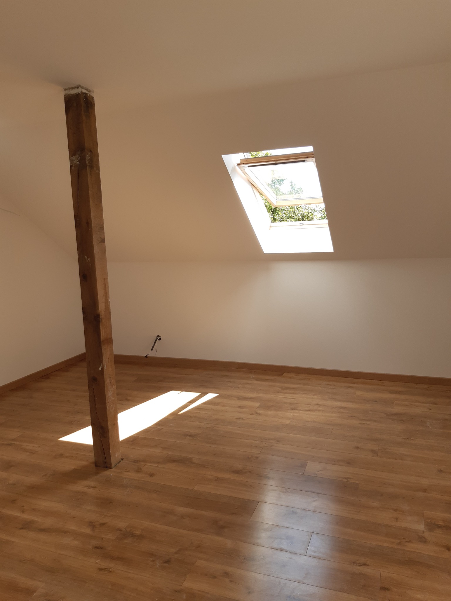 Poddasze z drewnianą podłogą, belką konstrukcyjną i oknem dachowym wpuszczającym światło. Widoczne gniazdko elektryczne na ścianie.